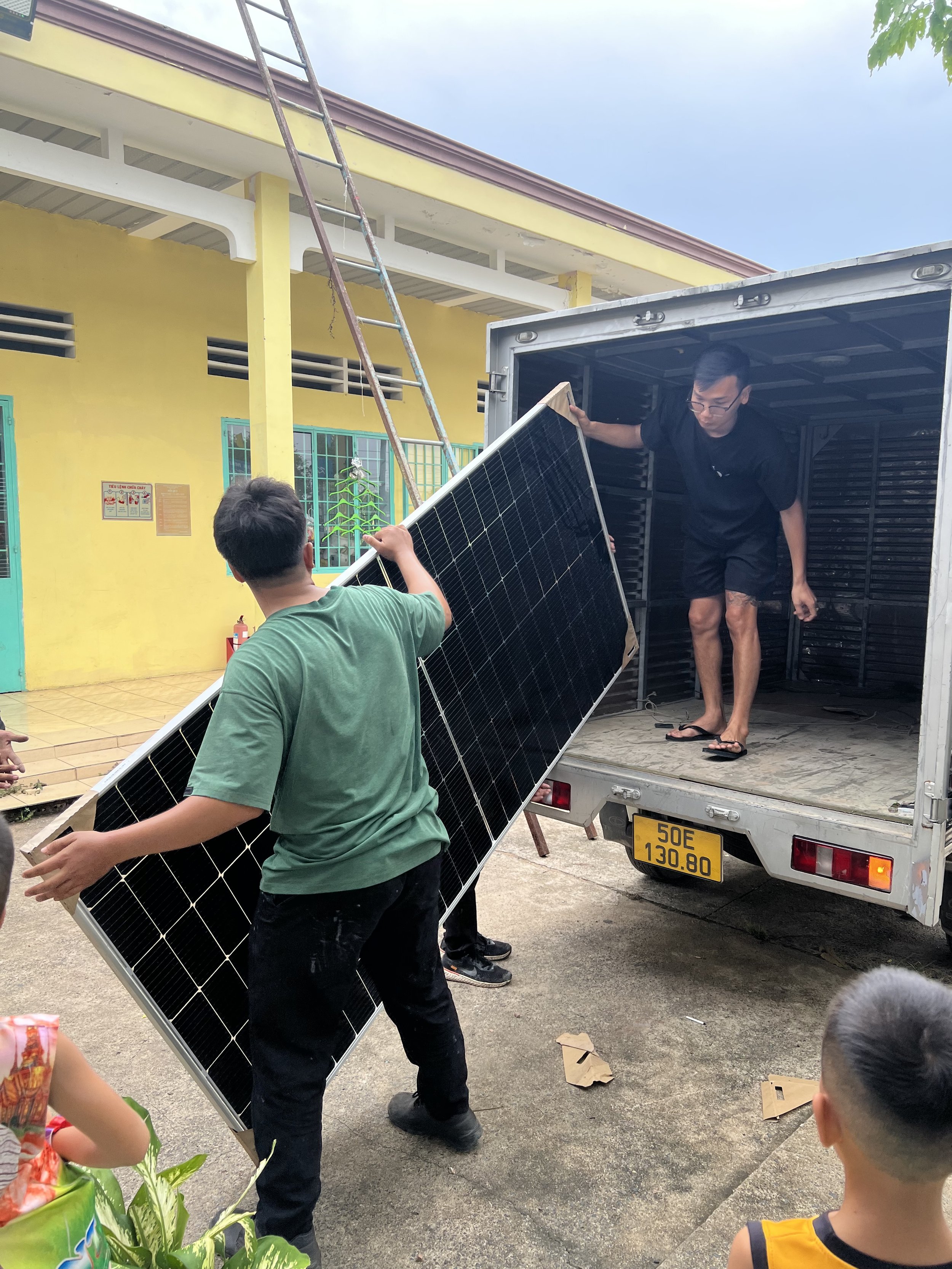 Workers unloading solar panels from a delivery truck at an orphanage, with children watching nearby