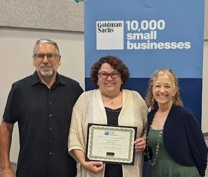 Three people standing in front of a banner that reads "Goldman Sachs 10,000 Small Businesses." The person in the middle is holding a framed certificate and smiling. The other two are also smiling.
