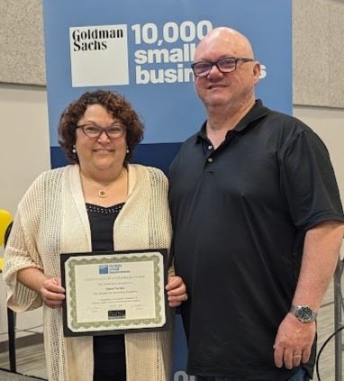 A woman holding a certificate standing next to a man in front of a blue Goldman Sachs banner that reads '10,000 small businesses'.