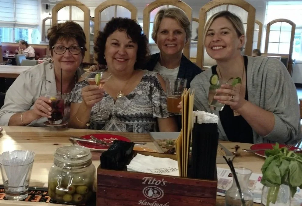 Four smiling women sitting at a restaurant table holding drinks, with food and condiments on the table behind them.