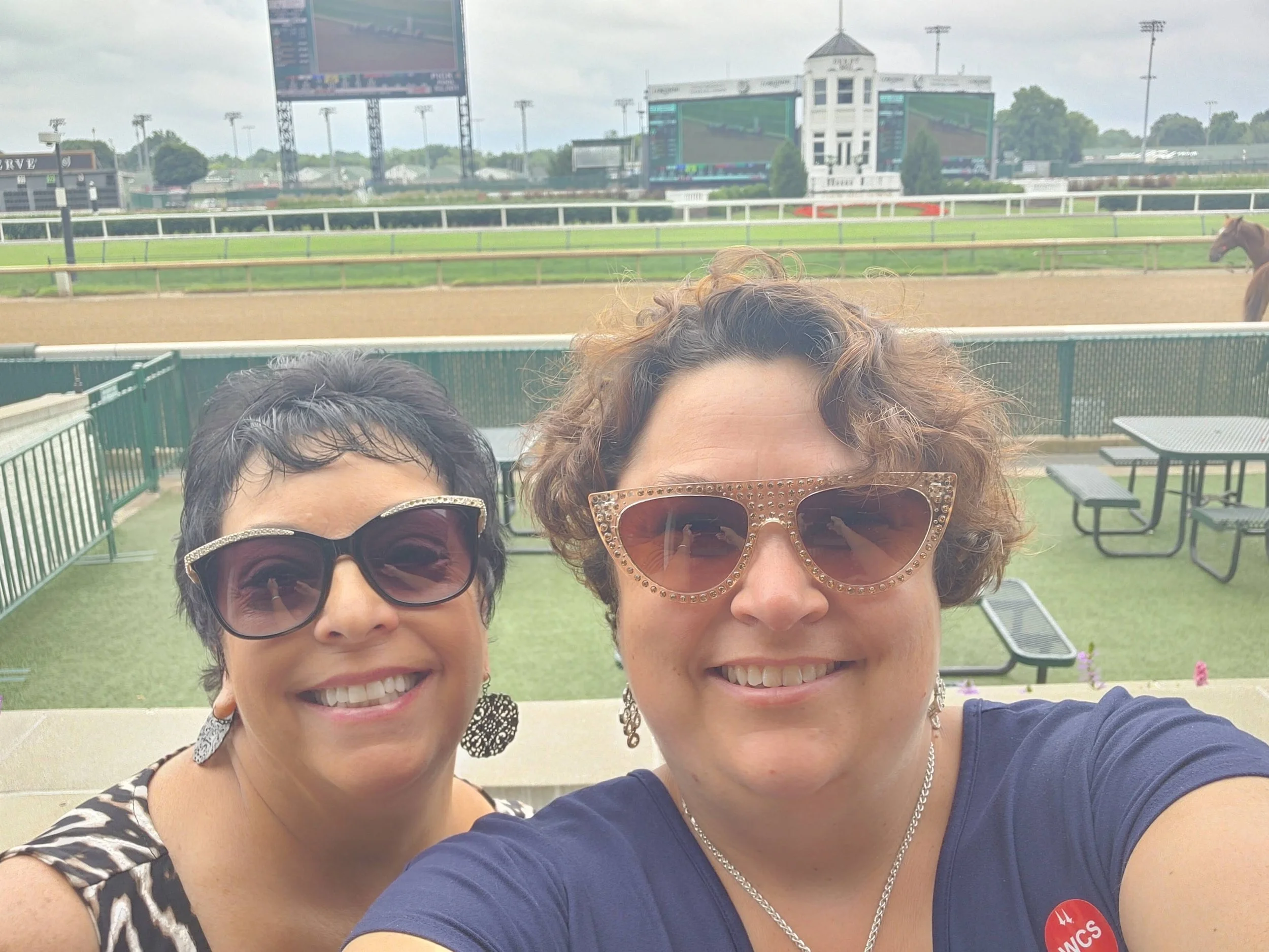 Two women wearing sunglasses smiling in front of a racetrack, with a large screen and horse in the background.
