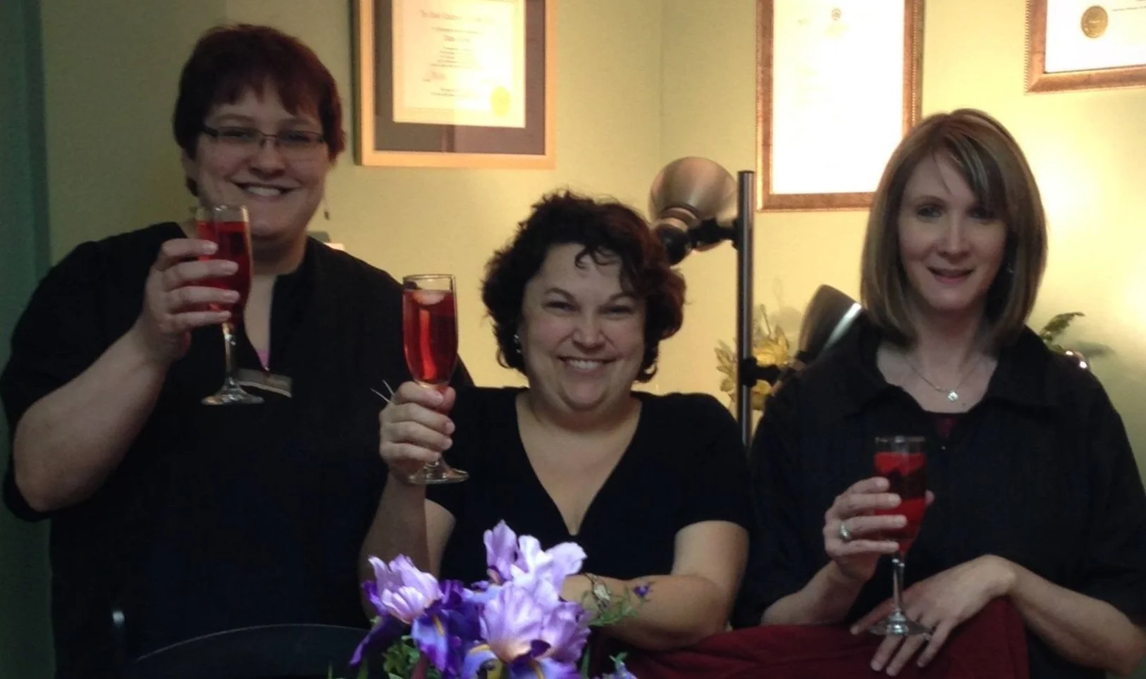 Three women smiling and holding glasses of red drink in a room decorated with framed certificates and flowers.