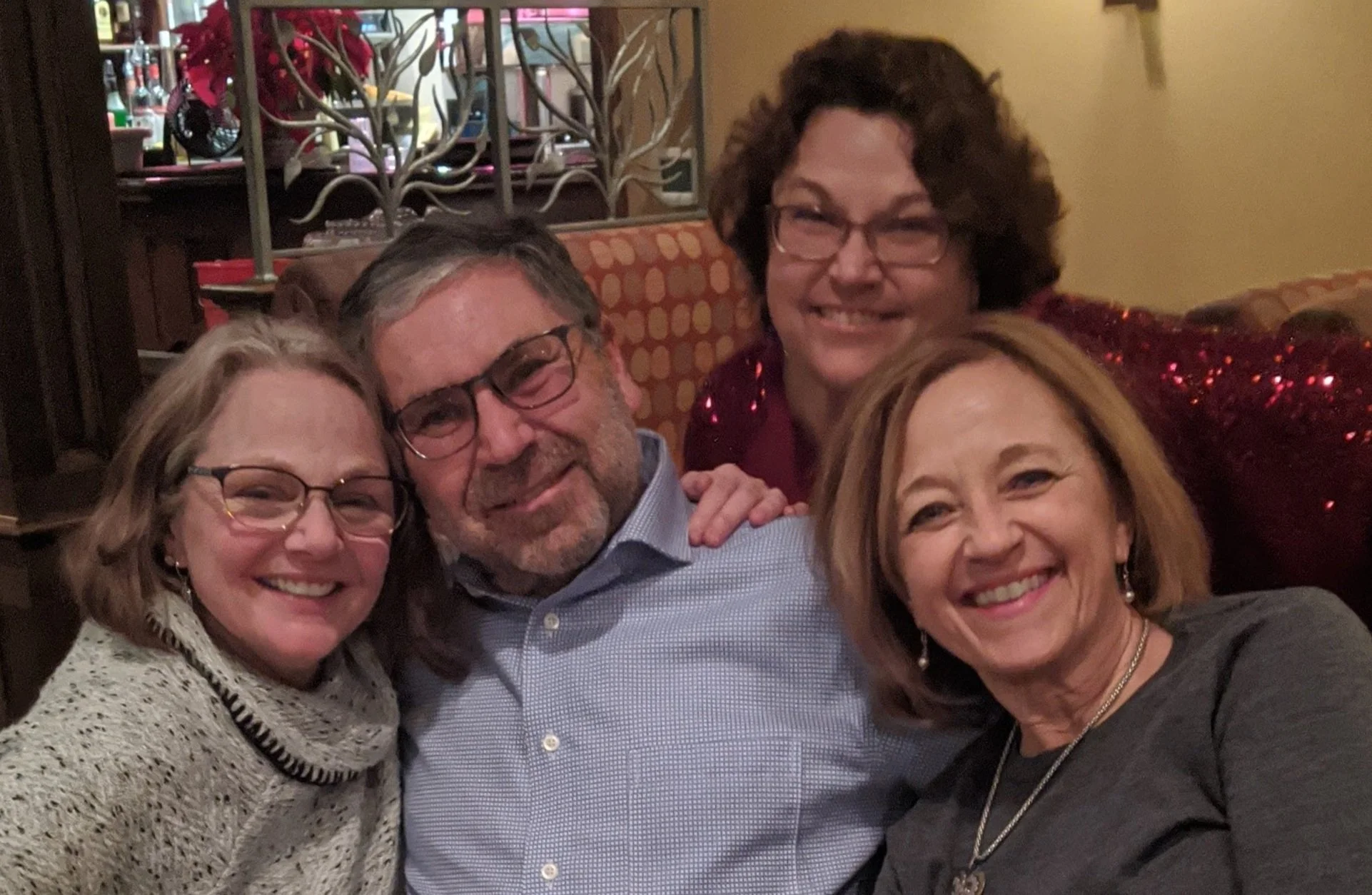 Four smiling adults posing together indoors, with a background of a restaurant or cozy setting.