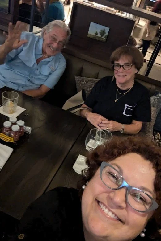 Three people taking a selfie at a restaurant table, smiling. Two women are sitting at the table and a man is behind them, waving. The table has drinks and condiments.