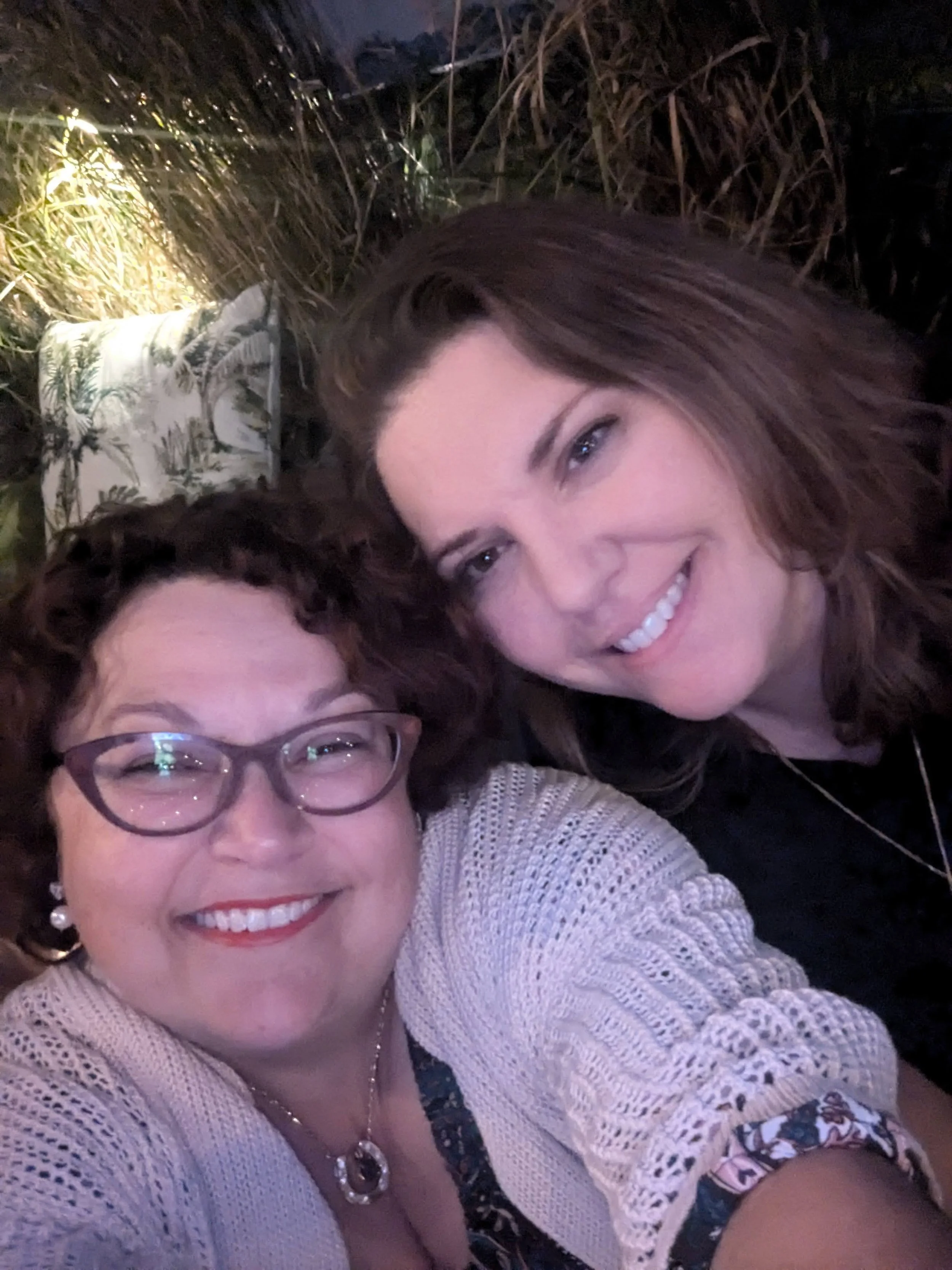 Two women smiling and taking a selfie together outdoors at night, with tall grass and plants in the background.