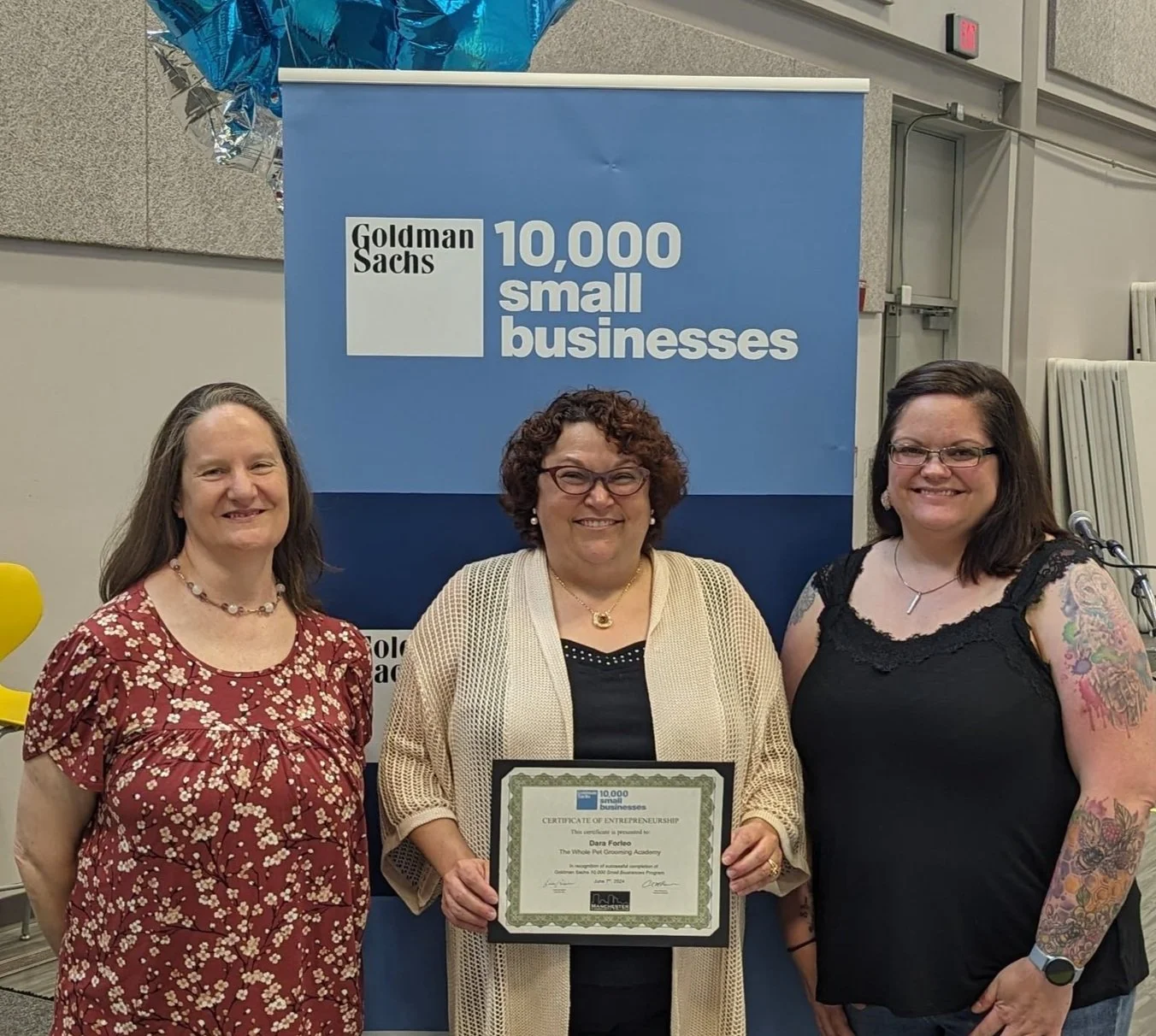 Three women standing in front of a blue Goldman Sachs banner that says '10,000 small businesses.' The woman in the middle holds a framed certificate.