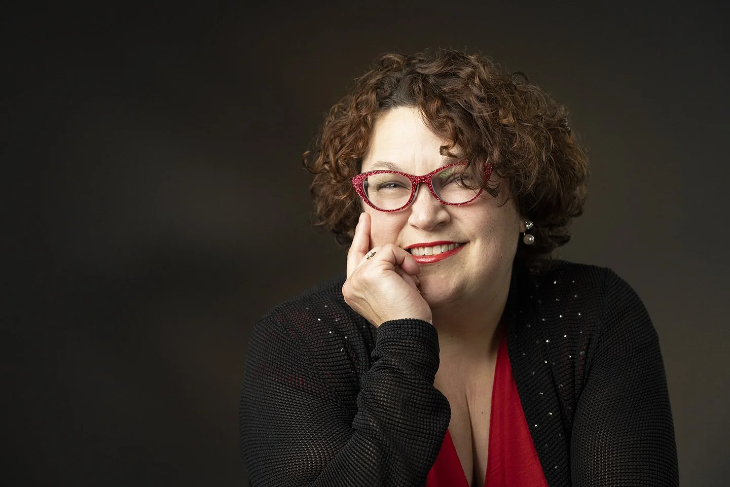 A woman with curly brown hair, wearing red glasses, a black jacket with sparkles, and a red top, smiling and resting her face on her hand, against a dark background.
