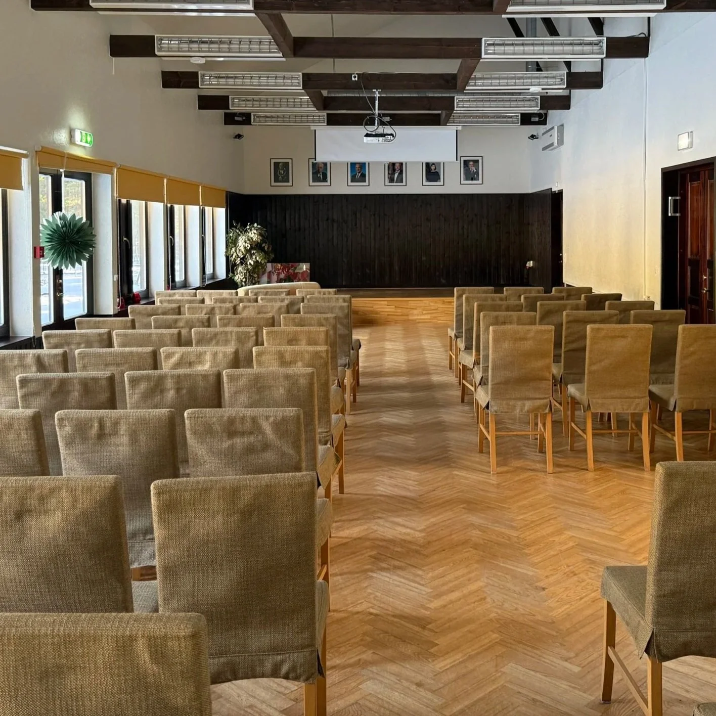 An empty conference room with rows of beige upholstered chairs facing a stage with a dark wooden backdrop. There is a large projection screen above the stage and framed portraits on the wall. The room has large windows along the side with yellow blinds, and the floor is wood-patterned.