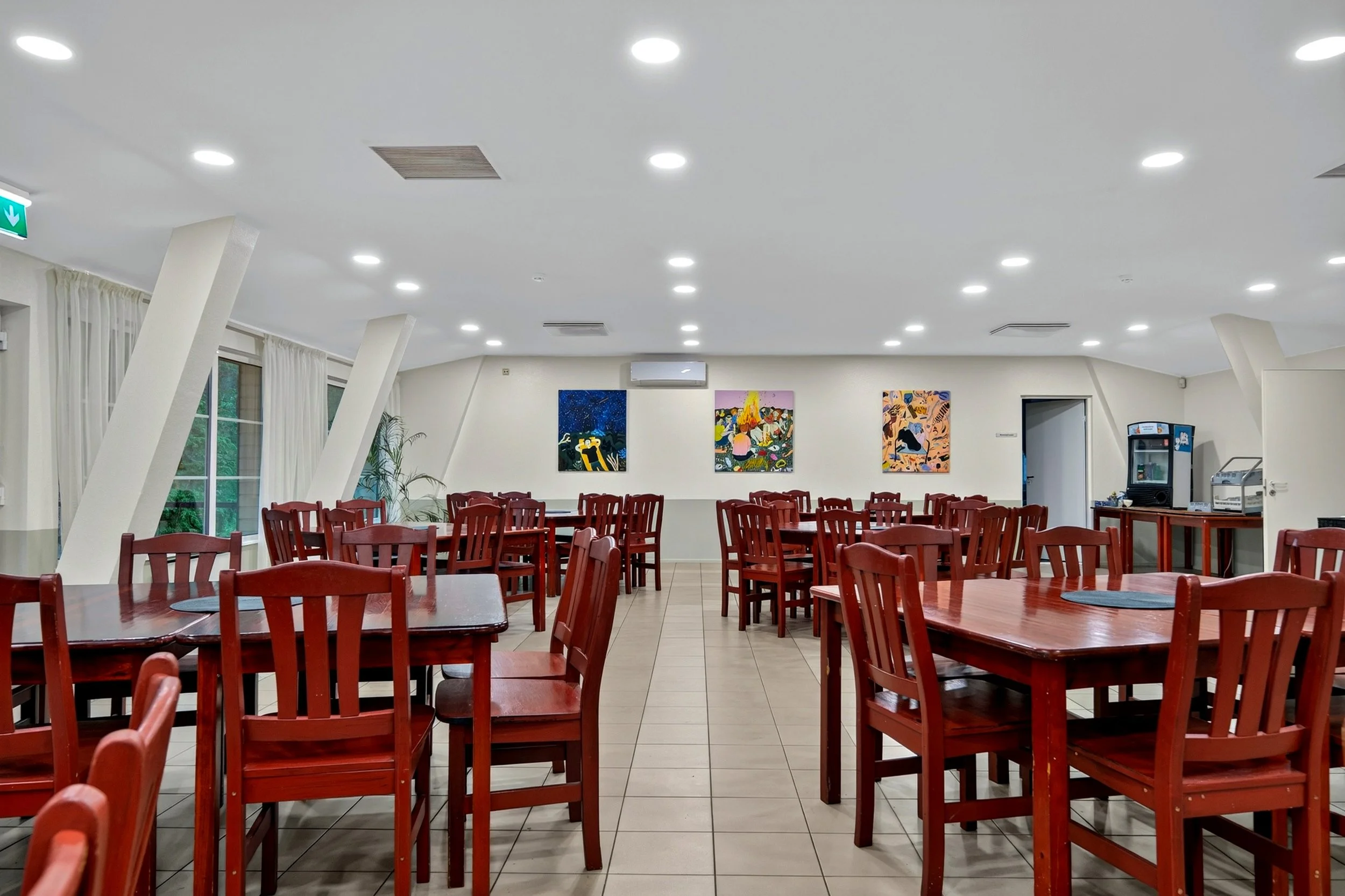 A dining area with red wooden chairs and rectangular tables, artwork on the walls, large windows with white curtains, ceiling lights, and an air conditioning unit.