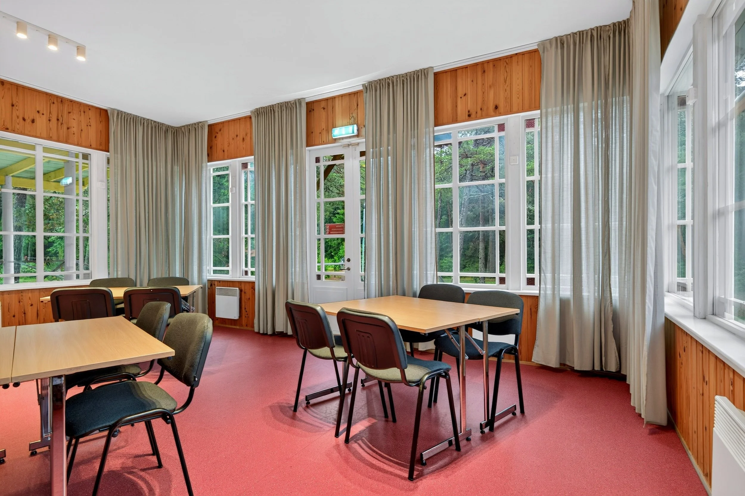 Interior of a room with several tables and chairs, large windows with beige curtains, wood-paneled walls, and a view of trees outside.