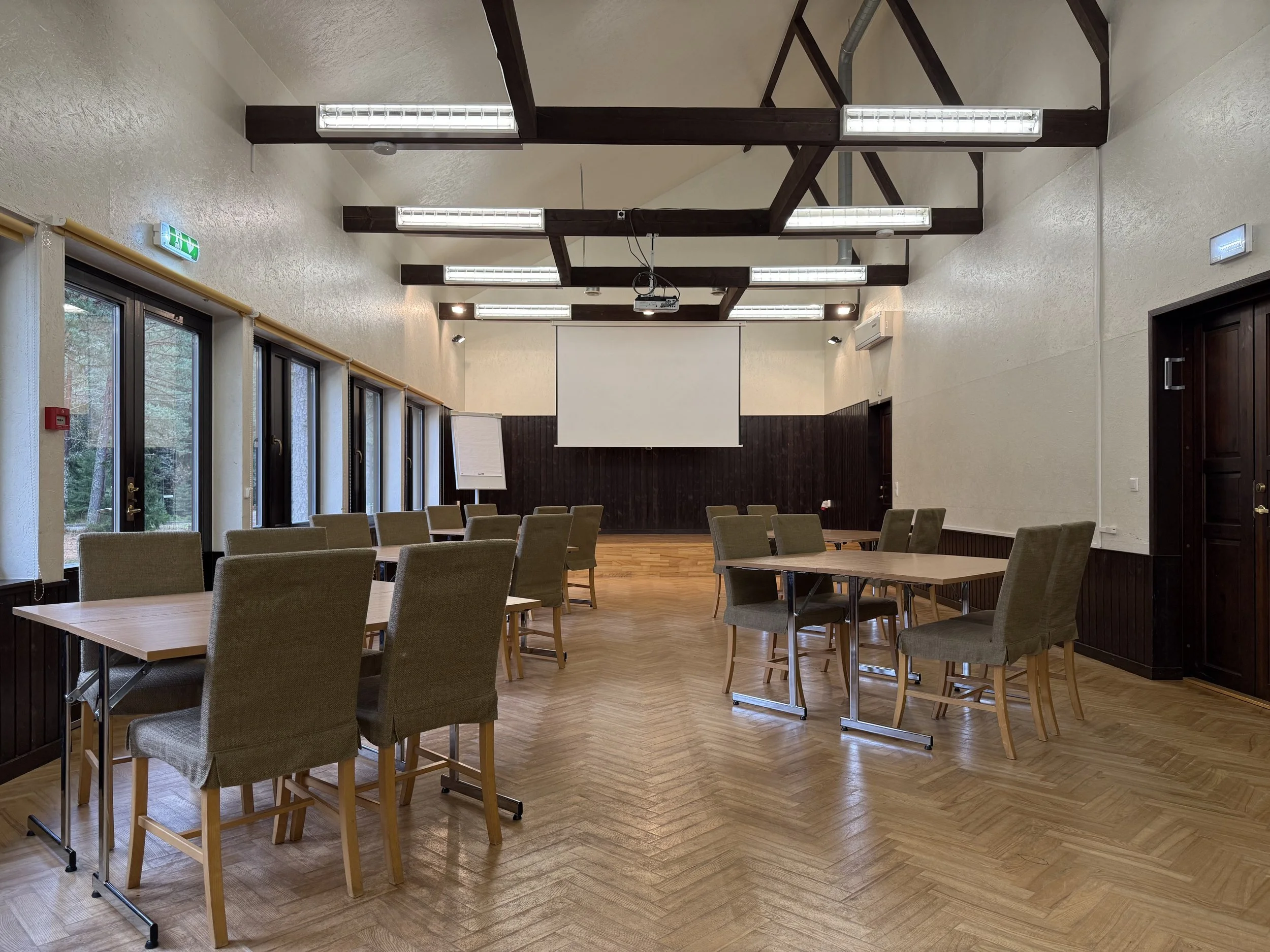 A conference room set up with tables and chairs, a large screen at the front, and ceiling-mounted fluorescent lights.