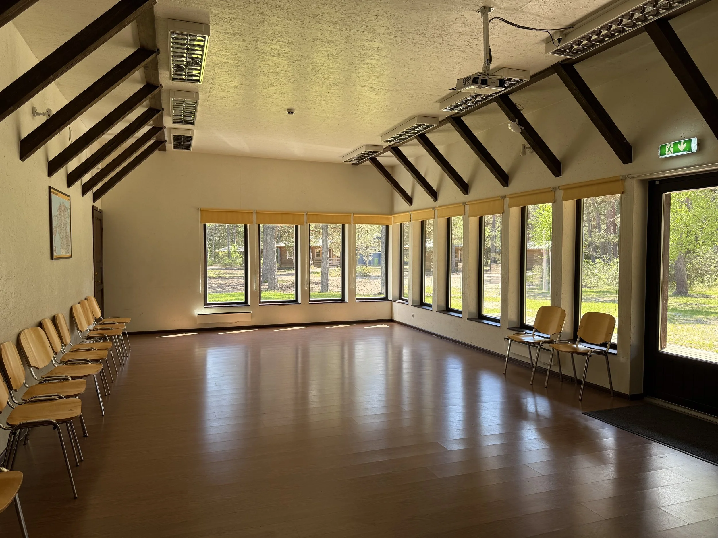 Empty room with wooden chairs arranged in a semi-circle along the walls, large windows with yellow blinds, ceiling lights, a projector, an emergency exit sign, and outdoor trees visible through the windows.