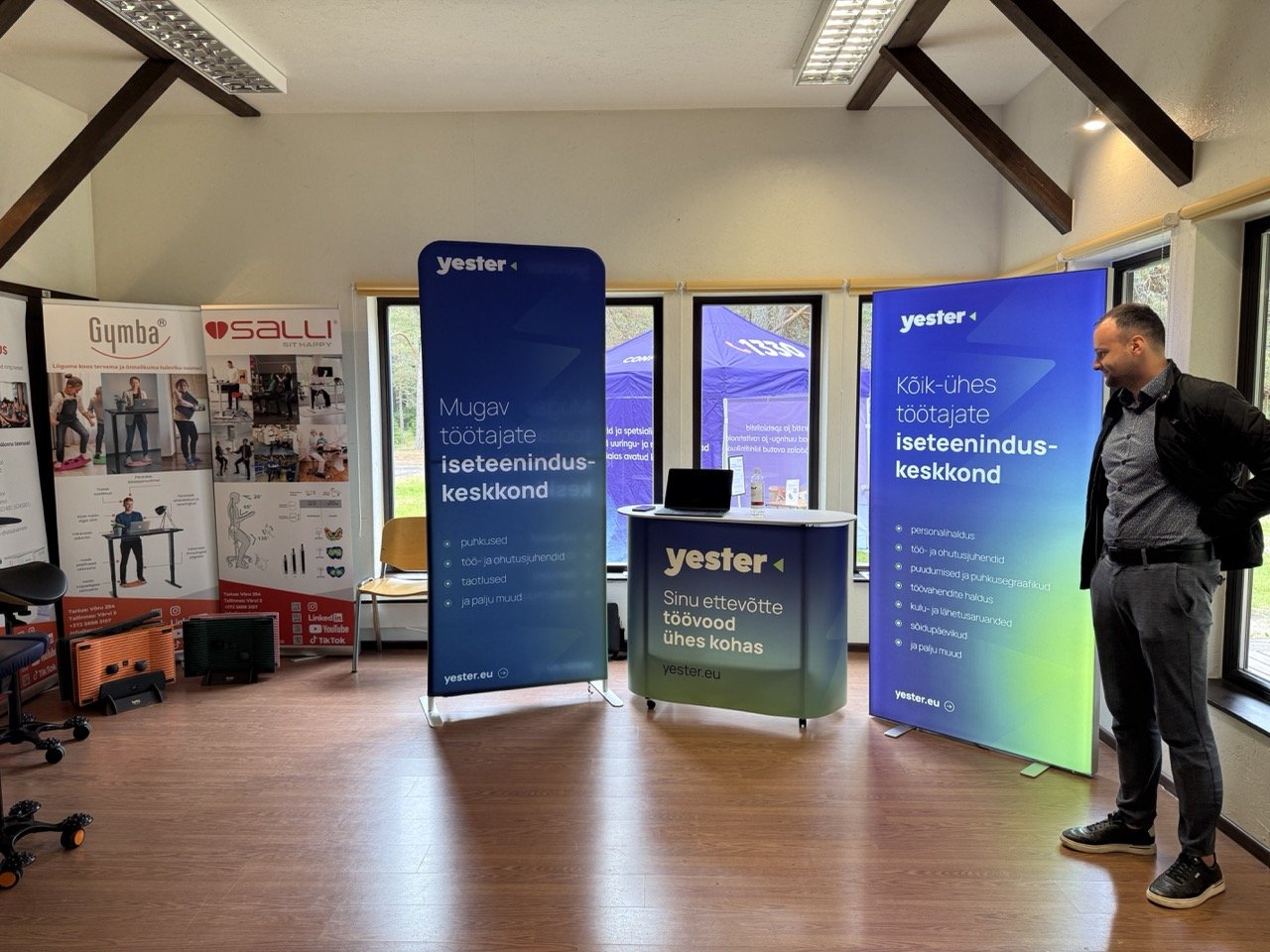 An indoor exhibition or conference room with display banners for Yester and Sali brands. Two large blue Yester banners promote the company's services, with a man standing near the right banner. Additional promotional materials and chairs are visible near the windows, which reveal a tent outside.