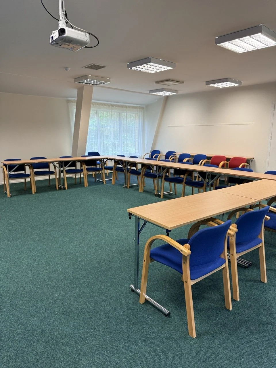 Empty conference room with U-shaped arrangement of tables and chairs, equipped with ceiling lights, a projector, and a window with sheer curtains.