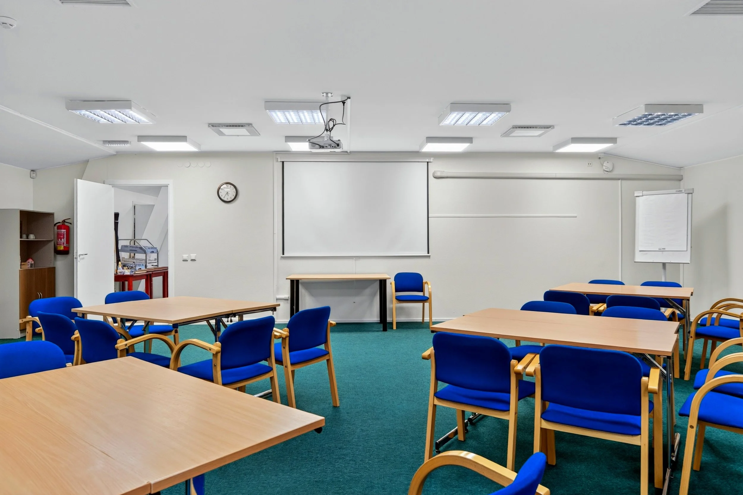 Empty classroom with blue chairs and wooden tables, white walls, a whiteboard, a clock, and ceiling lights.