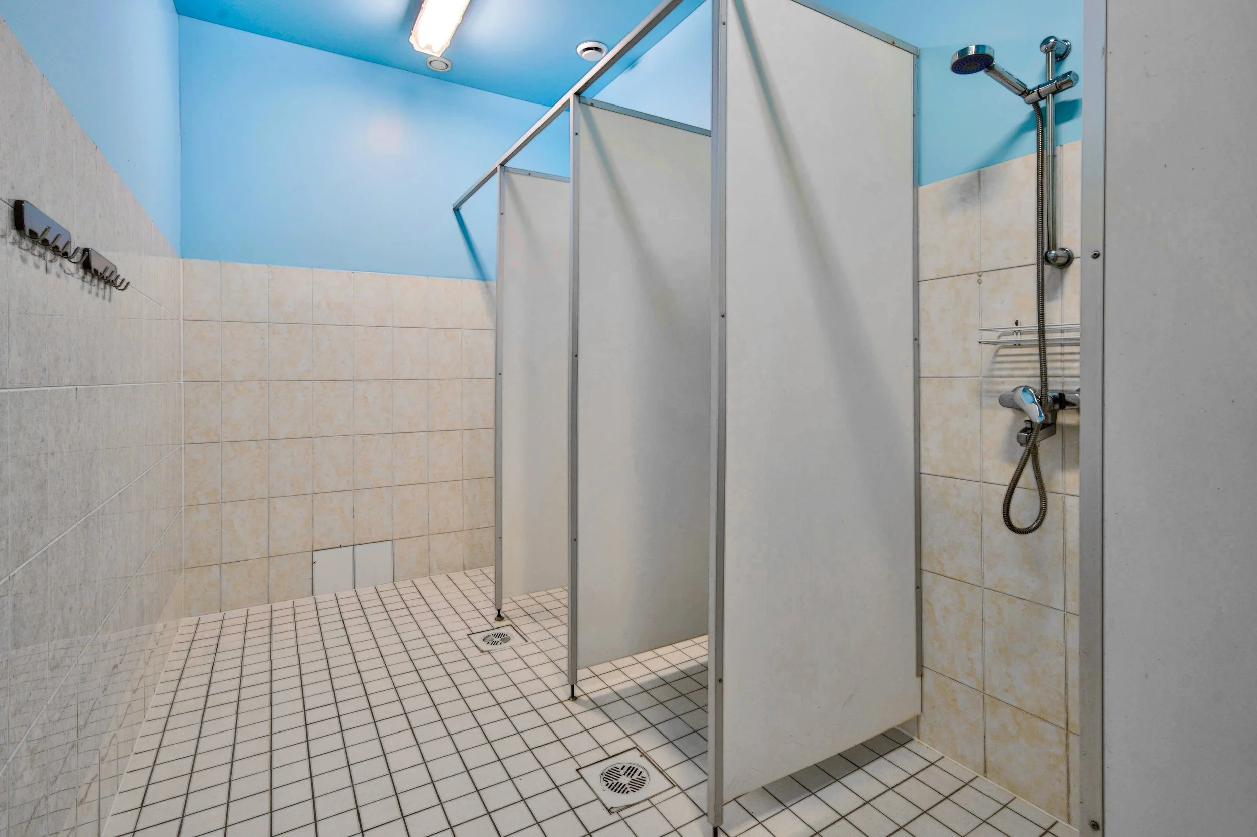 Public shower room with blue walls, white tiled partitions, and a handheld shower head.