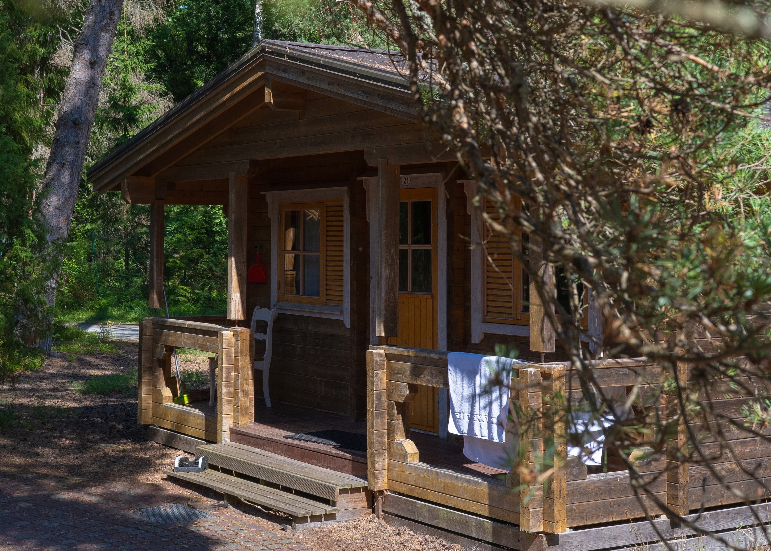 A small wooden cabin in a forested area with green trees surrounding it and a porch with towels hanging on the railing.