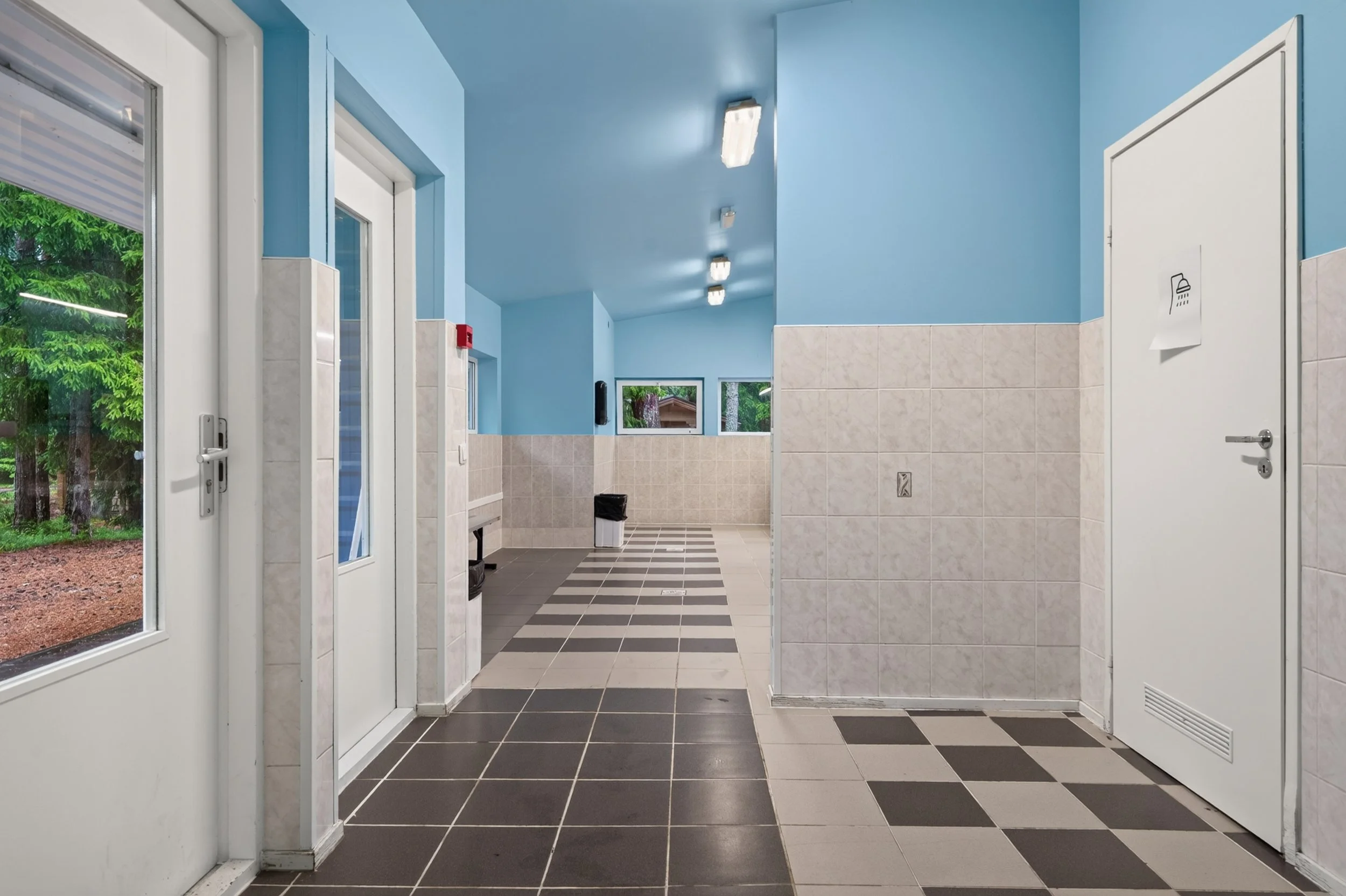 Public restroom with blue walls and black and gray checkered floor, featuring a shower with a sign, windows, and a glass door leading outside.