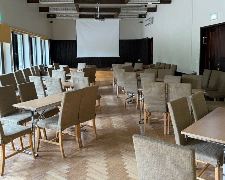 Empty conference room with rows of chairs and tables, a large blank screen at the front, and windows on the side.
