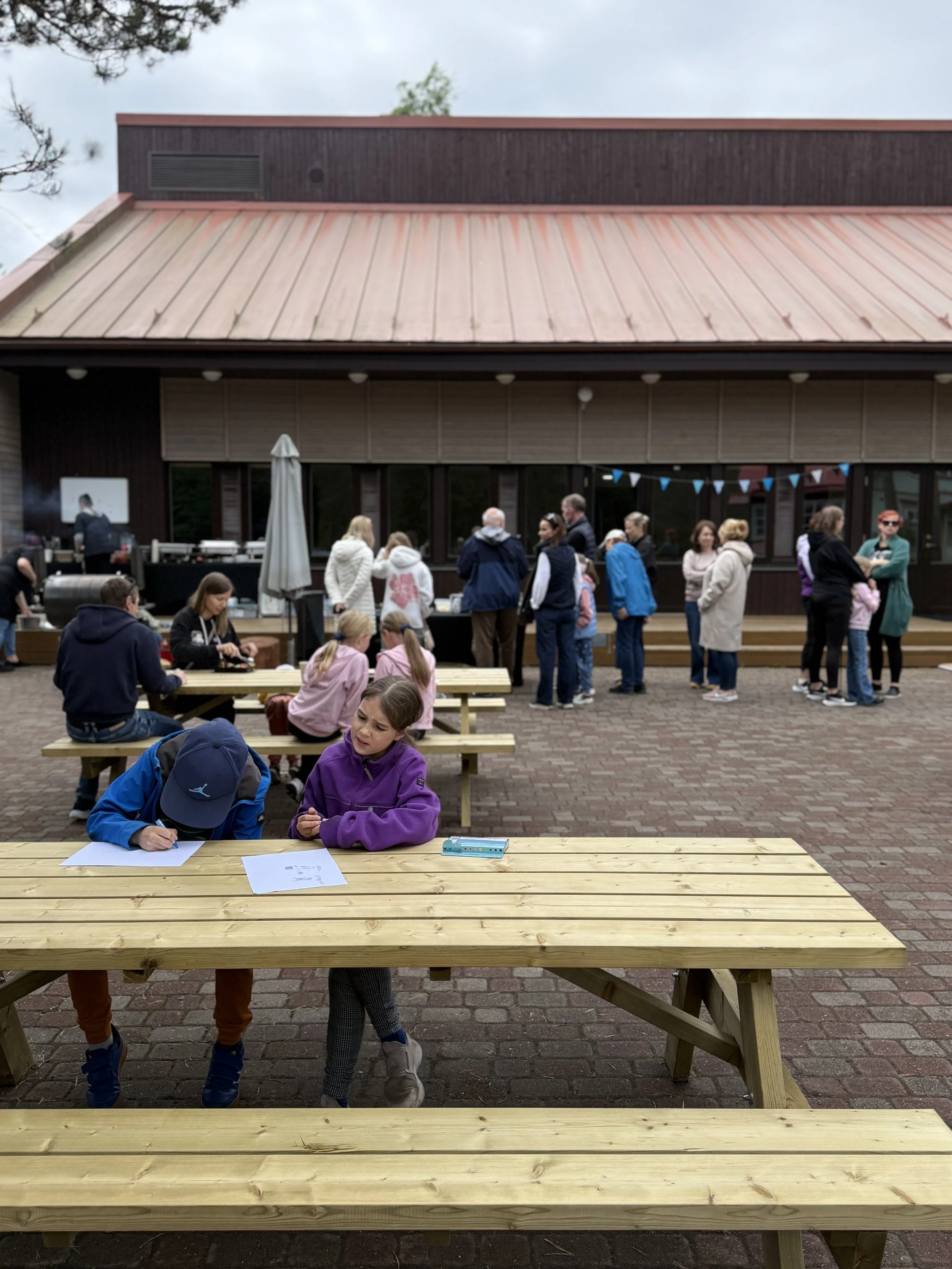 Children and adults gathering outdoors around wooden picnic tables, some sitting and others standing in line, with a building and cloudy sky in the background.