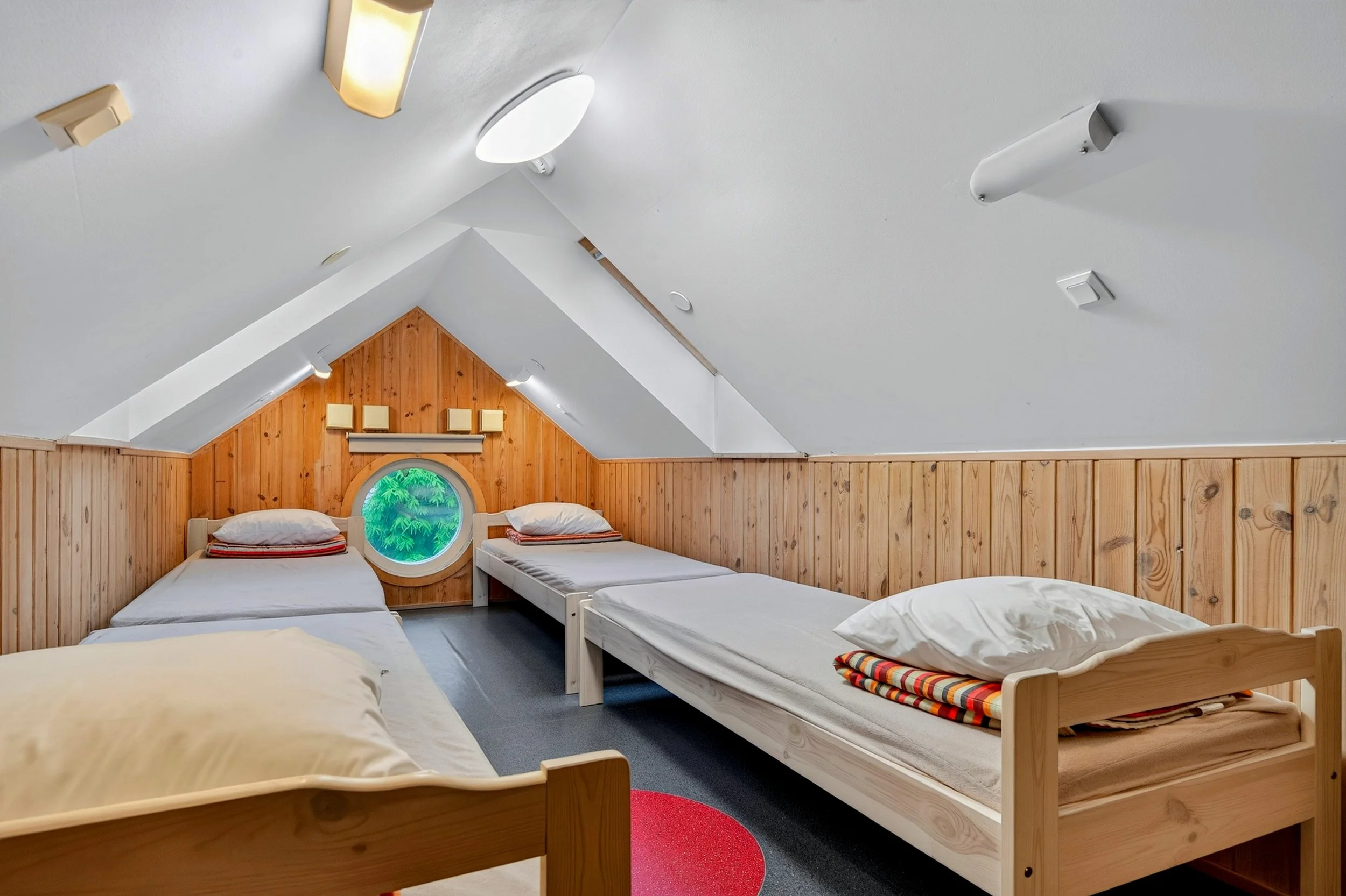 A room with a slanted ceiling containing four beds with wooden frames and white bedding. There is a small round window with a view of greenery, wooden paneling on the wall, and various ceiling lights and fixtures.