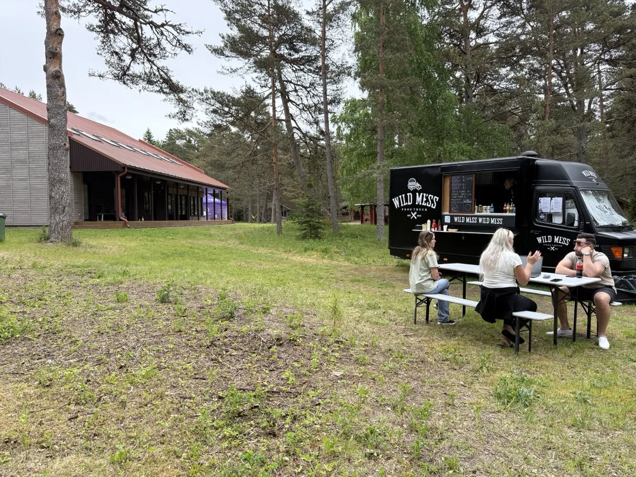 People sitting at a picnic table in front of a black food truck named 'Wild Mess' in a grassy outdoor area surrounded by trees.
