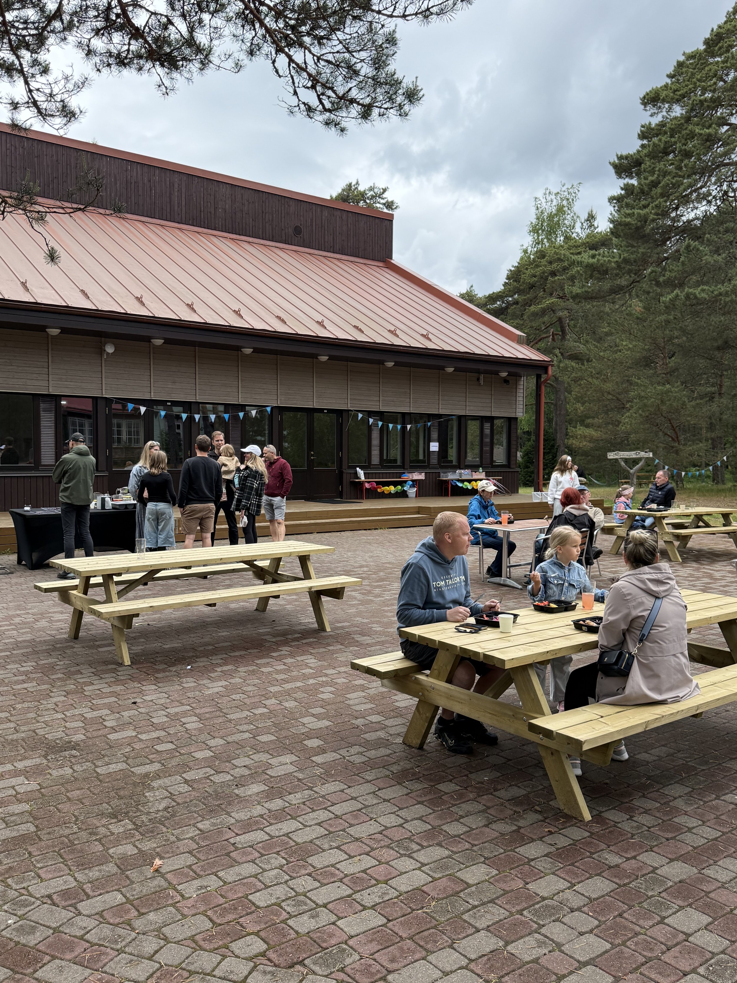 People gathered outdoors at a daytime event with picnic tables, a building with a metal roof, and trees in the background during overcast weather.