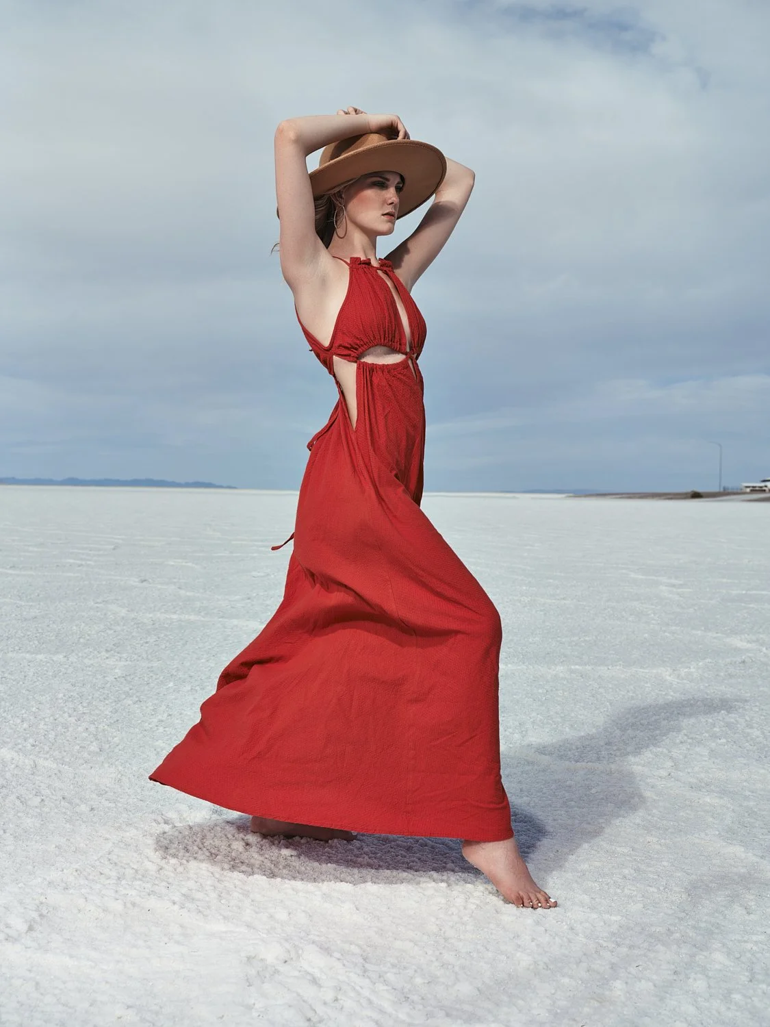A woman wearing a red dress and a wide-brimmed hat standing barefoot on a flat, white salt flats with a partly cloudy sky in the background.