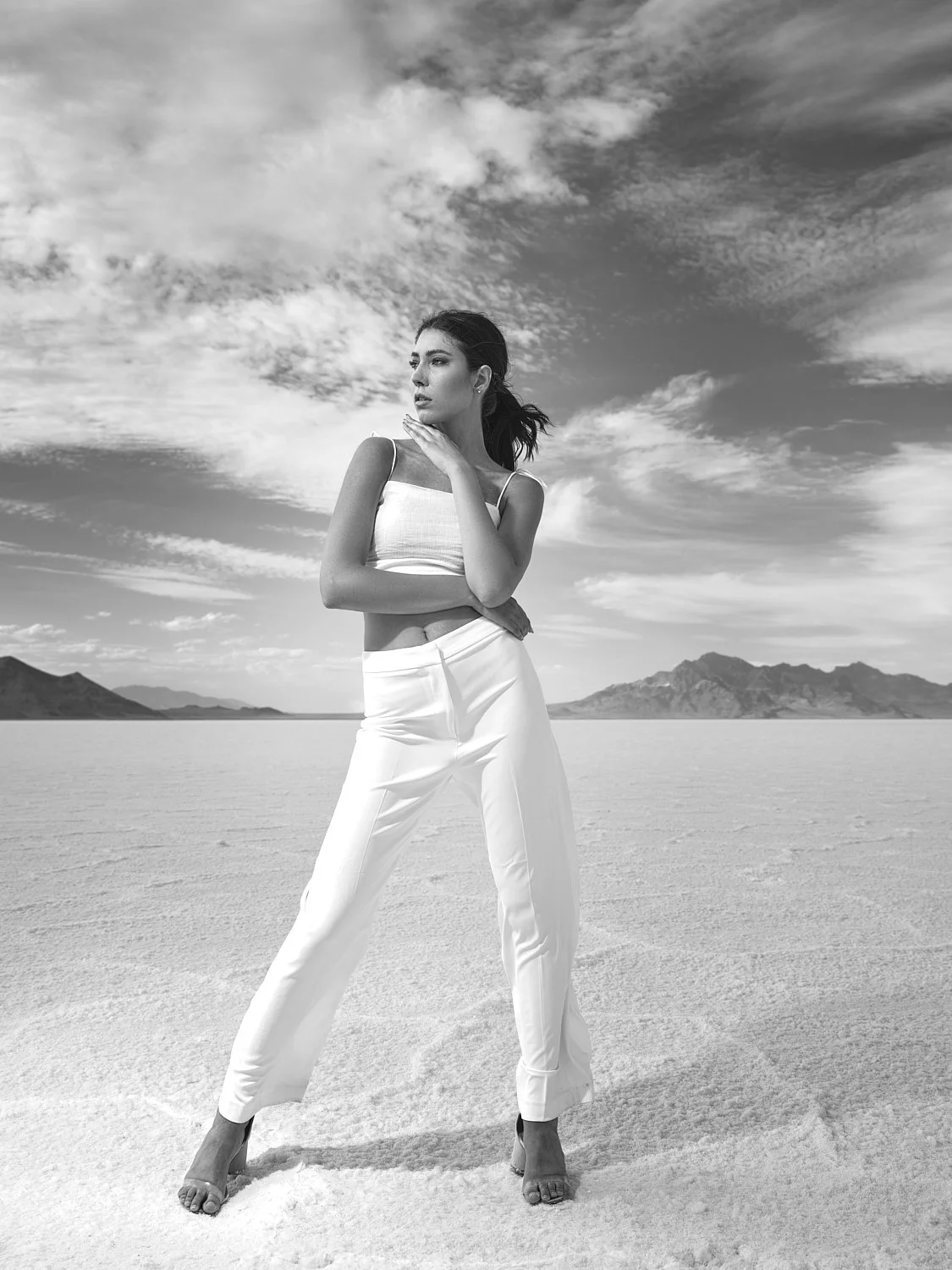 A woman standing on a salt flat, looking to the side with mountains in the background and a cloudy sky, wearing white pants and a sleeveless top.