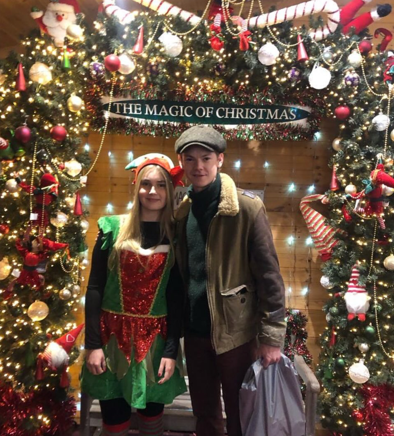 Thomas Brodie-Sangster with a young girl dressed as Santa's elf, in front of a Christmas archway and The Magic of Christmas sign