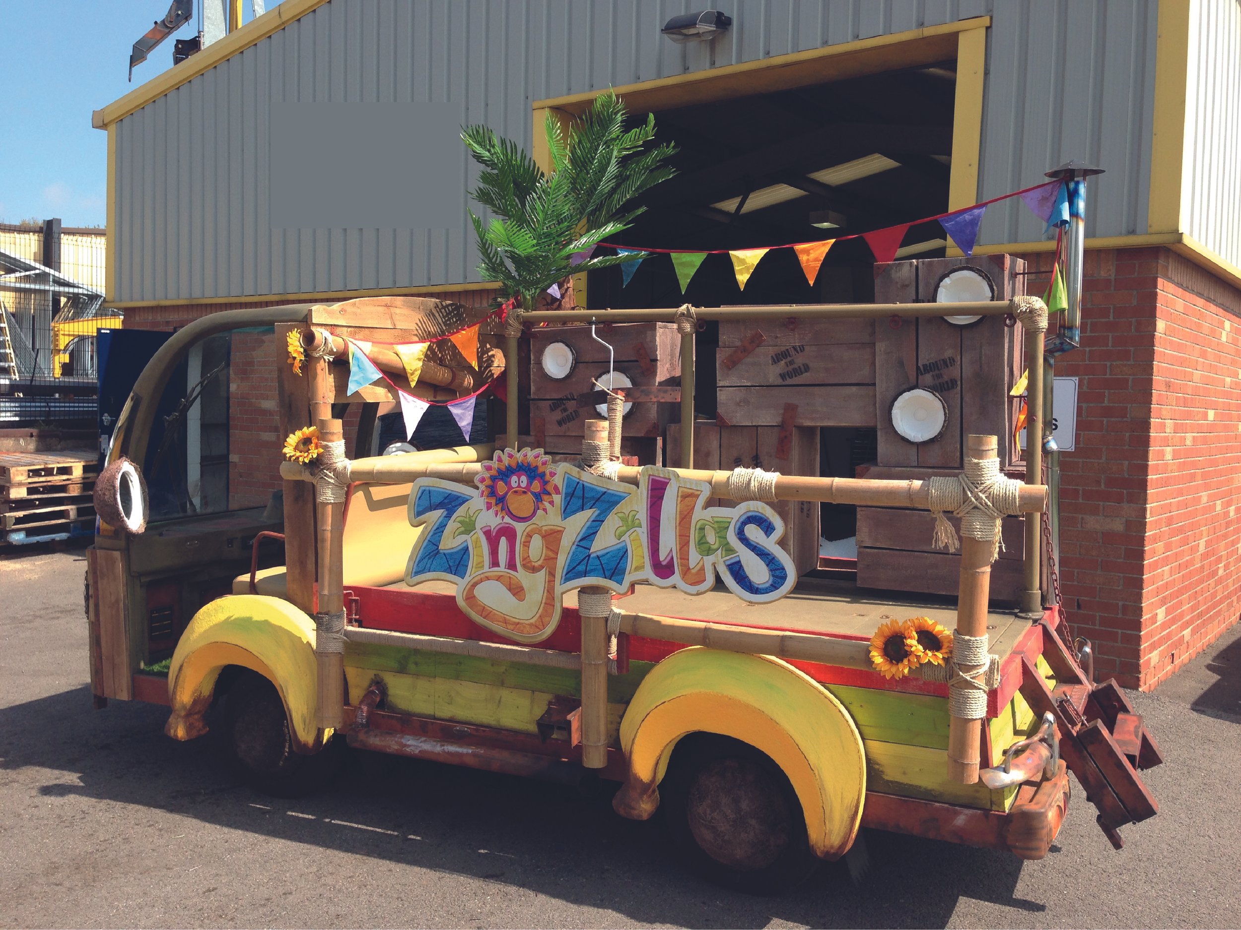 Colorful decorated vehicle with "ZingZillas" sign, featuring tropical design elements, bunting, wooden crates, and sunflowers, parked outside a building.