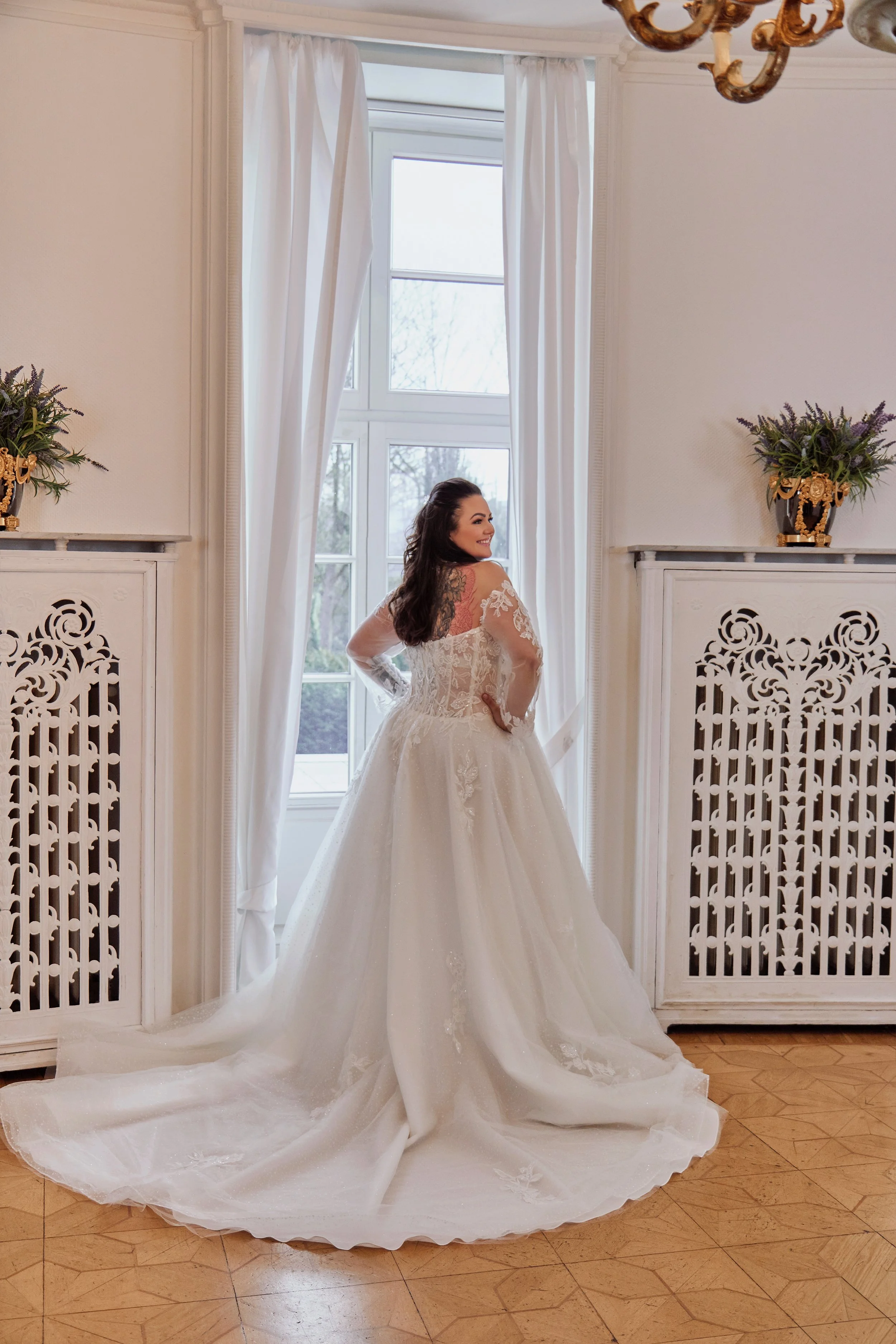 Back view of Lia Bella Model Tulla showing the illusion lace bodice, transparent long sleeves and wide glitter-tulle chapel train.