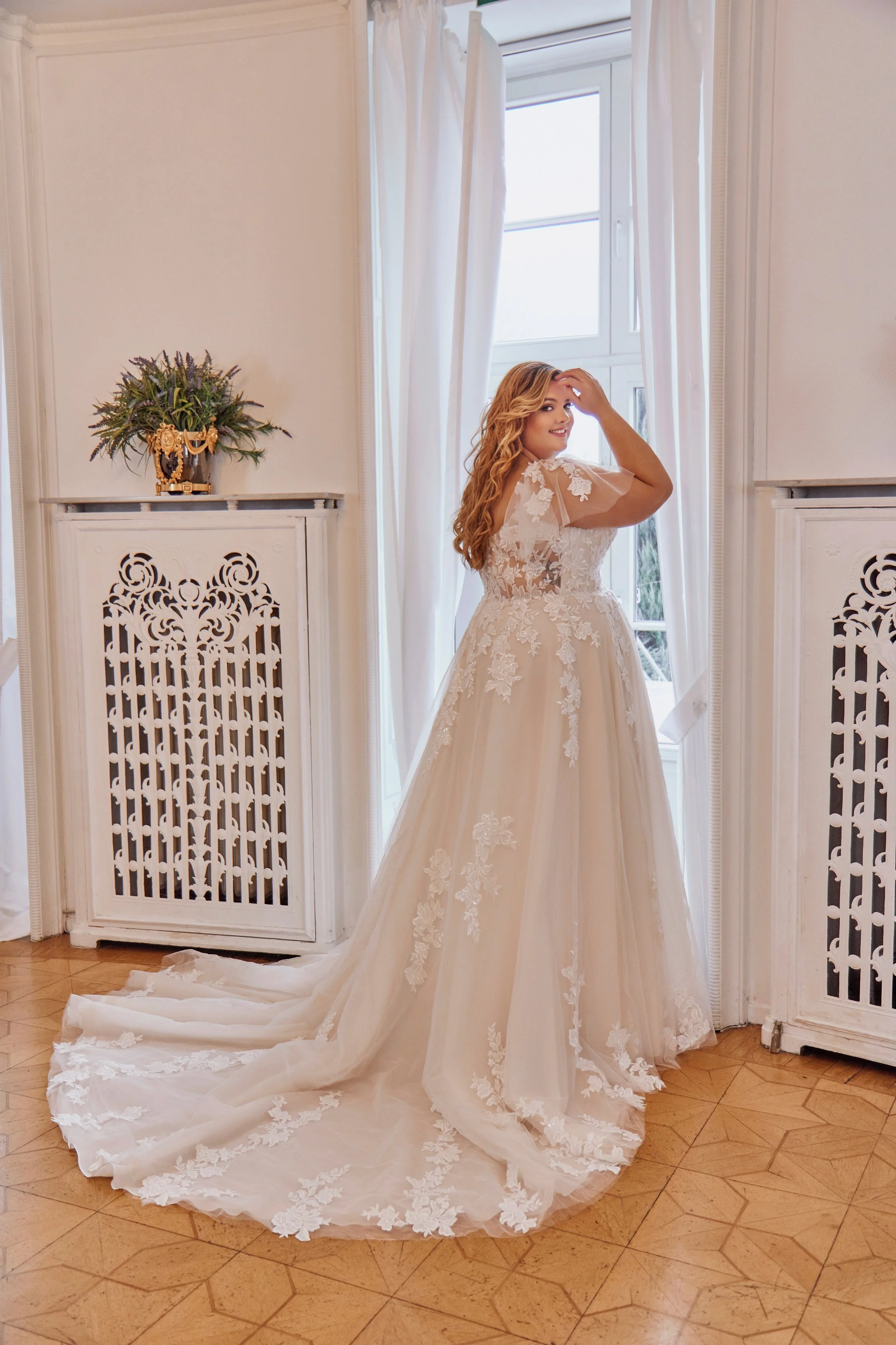 Three-quarter back view of Lia Bella Model Tasia showing the illusion lace bodice, flutter sleeves and floral appliqués along the train.