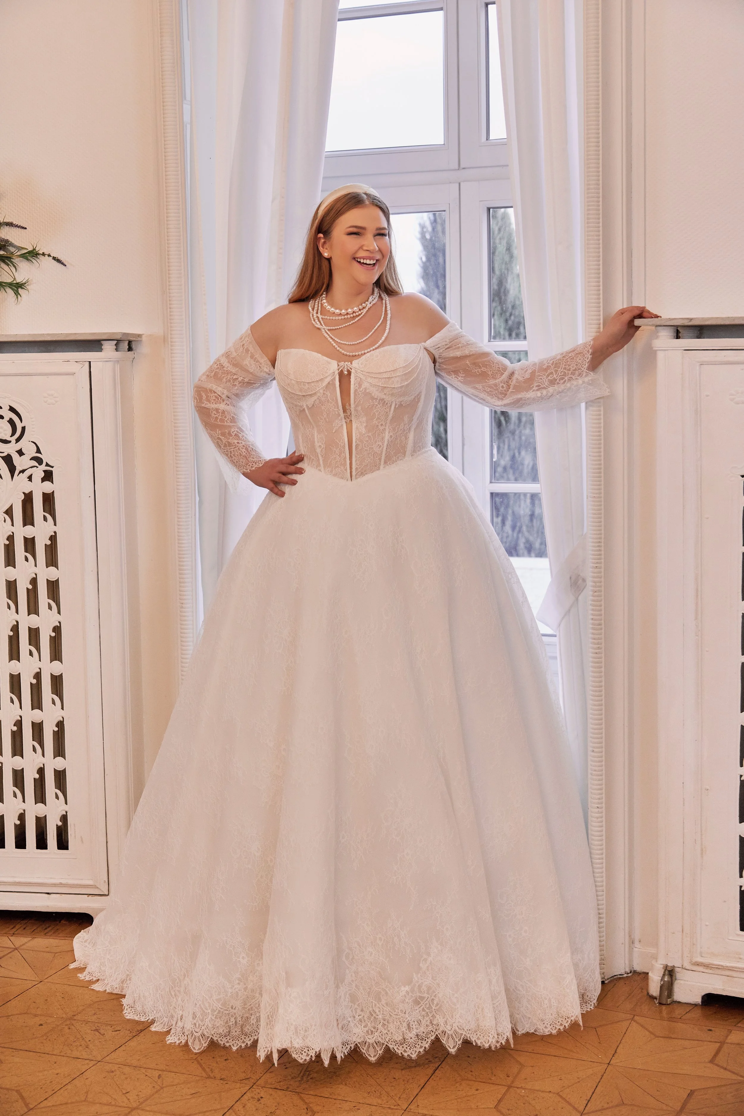 Full front view of Lia Bella Model Tia, an off-the-shoulder curvy lace ball gown with a structured illusion corset and long sleeves.