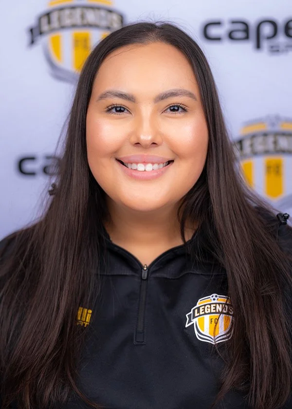A young woman with long brown hair smiling at the camera, wearing a black sports jacket with a 'Legends FC' logo, standing in front of a backdrop with logos including 'Legends' and 'cape'.