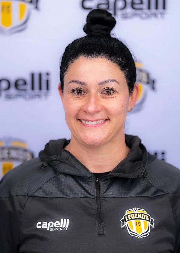 A woman with dark hair in a top bun, smiling, wearing a black sports jacket with a 'Legends FC' logo and 'Capelli Sport' logo, standing in front of a backdrop with similar logos.