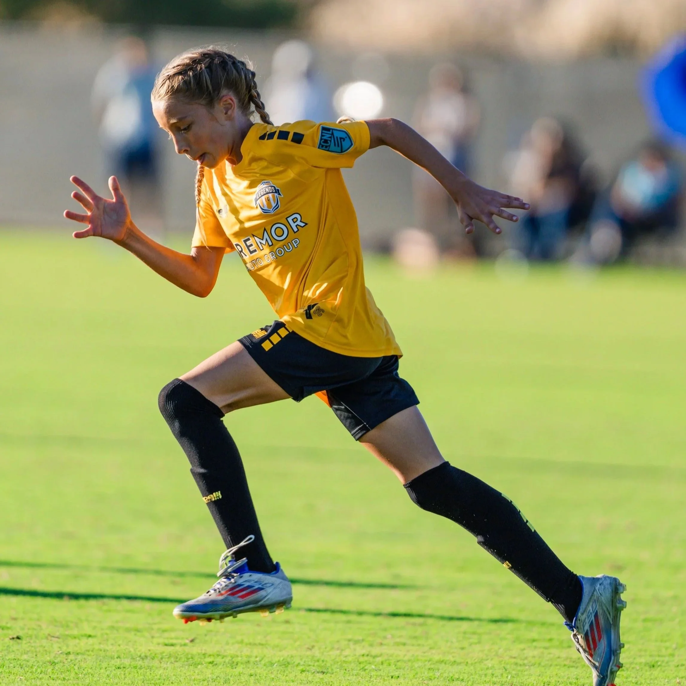 A female soccer player in a yellow jersey and black shorts running on a grassy field during a match.