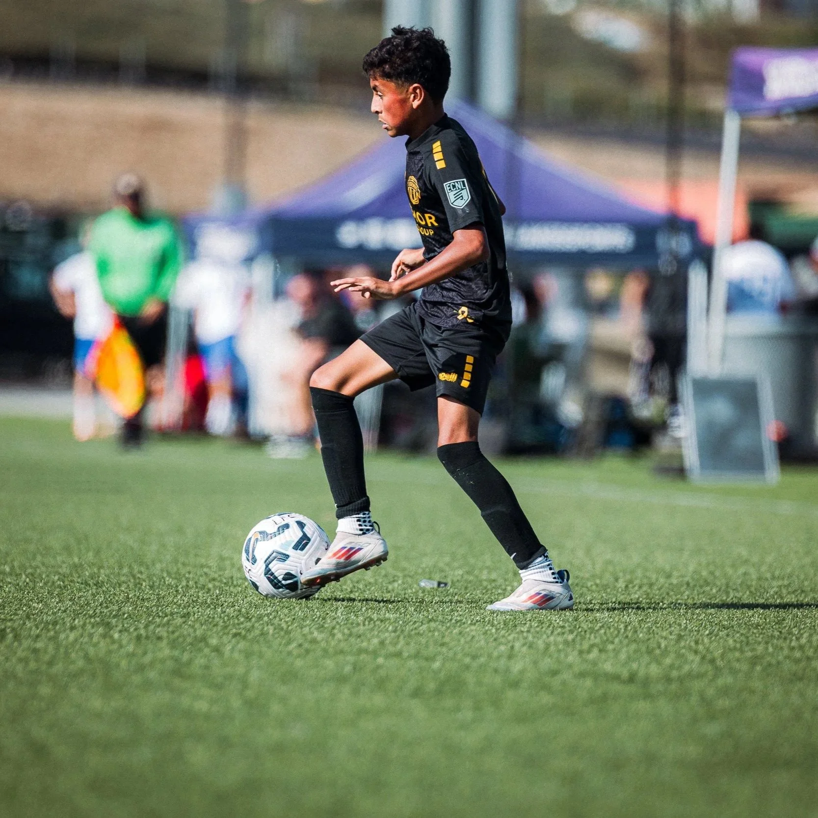 A young soccer player in black uniform and cleats controlling a soccer ball on a grassy field during a game.