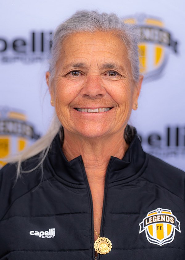 A smiling elderly woman with gray hair tied back, wearing a black jacket with a logo that says Legends FC, standing in front of a backdrop with the same logo and a blurred background.