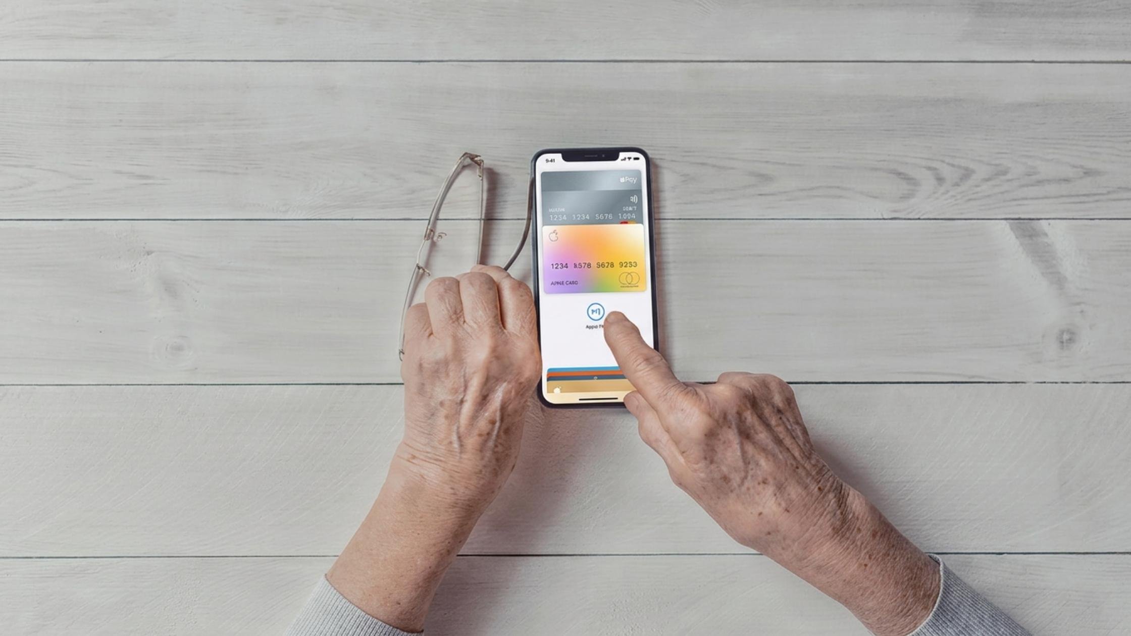 Older hands using a smartphone to access a digital wallet, with glasses held in the other hand