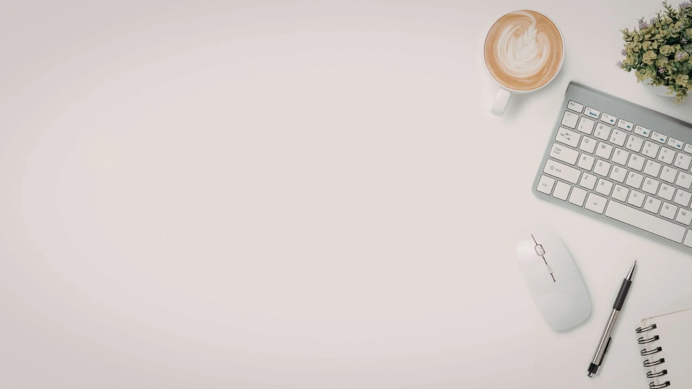 Minimalist workspace flatlay with a keyboard, notebook, pen, coffee and plant on a clean white surface.