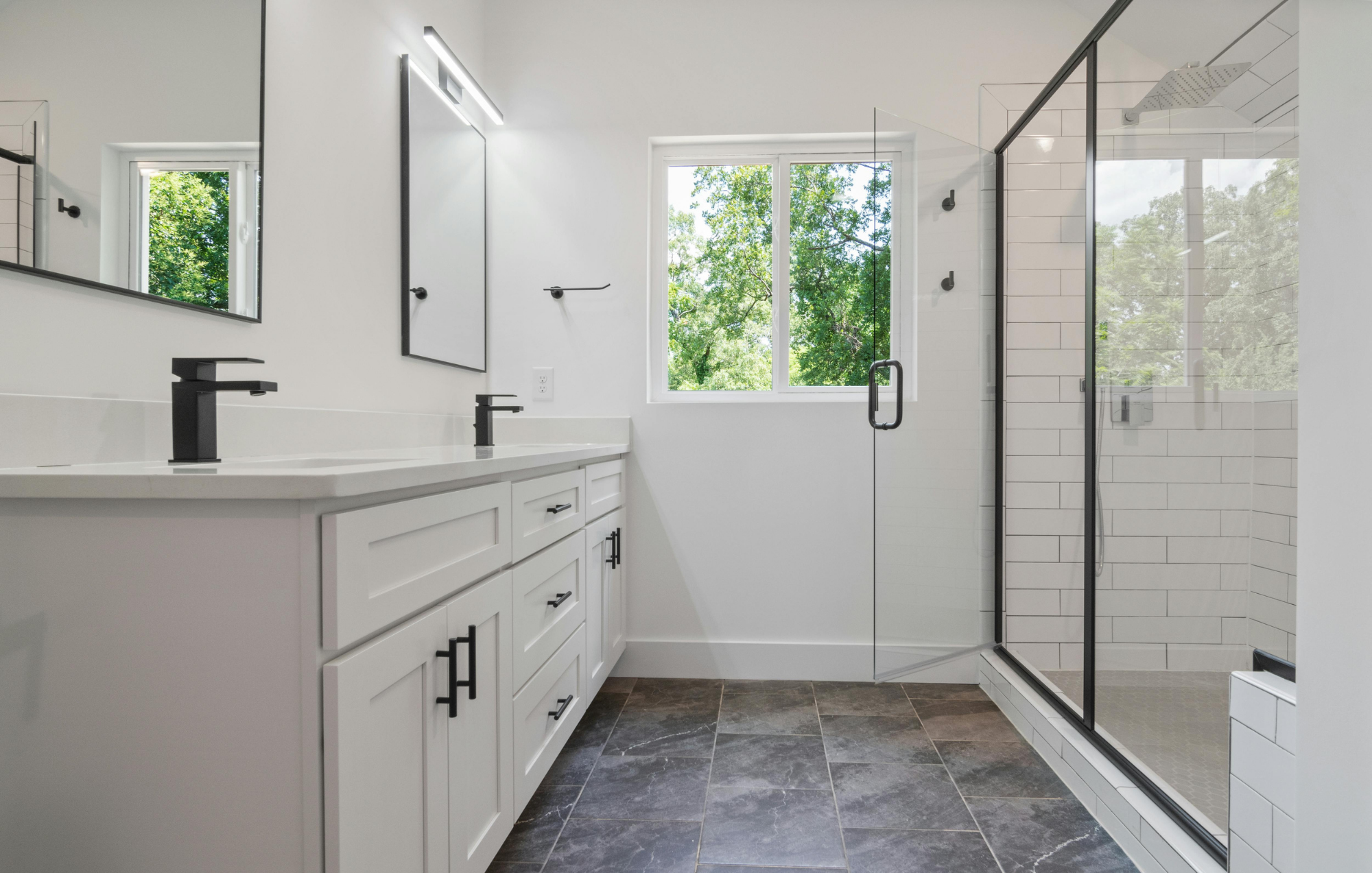 Modern bathroom with white vanity, black fixtures, large mirror, window showing green trees, and a walk-in shower with glass door and white subway tile