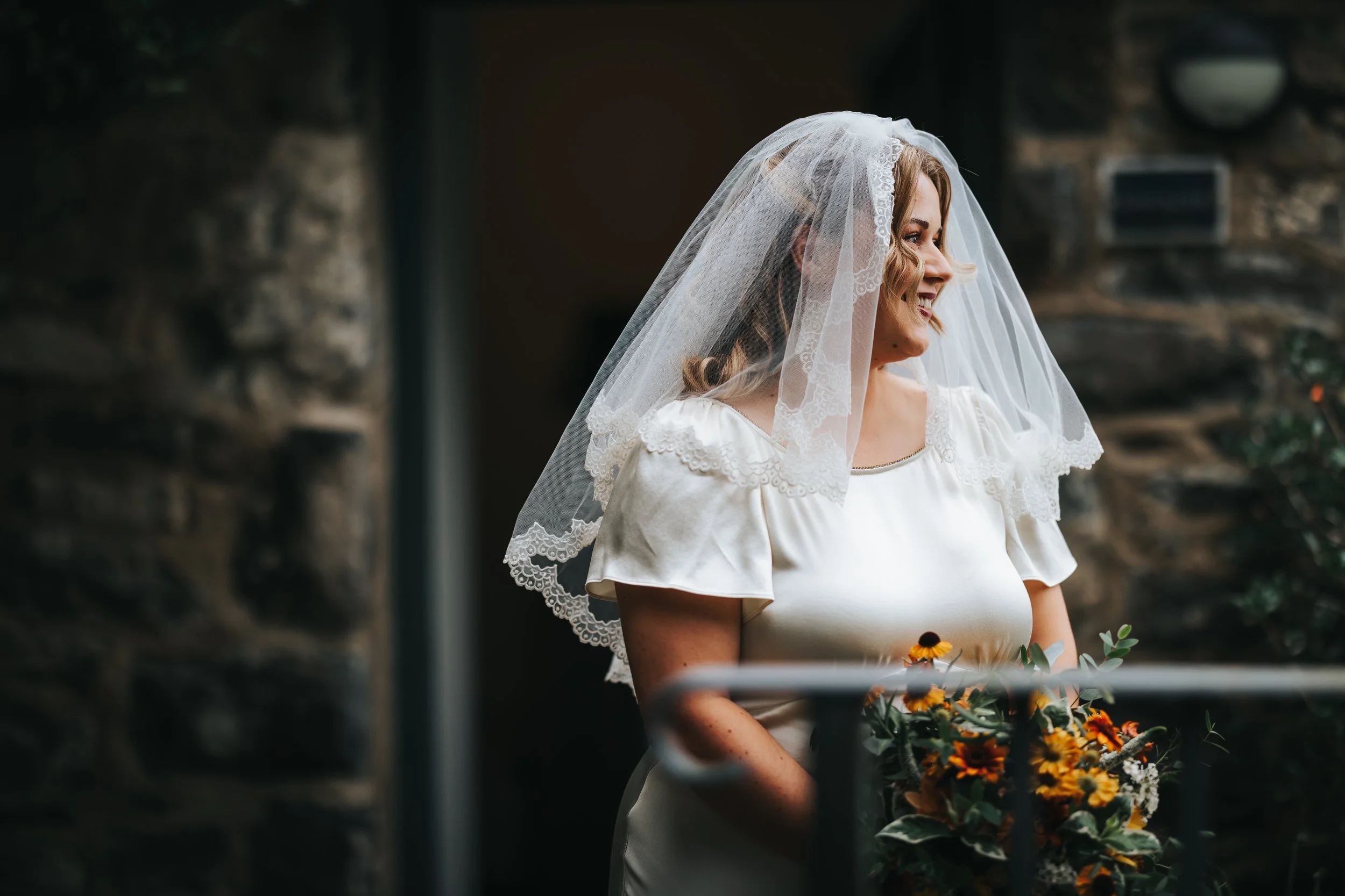 A woman dressed as a bride, wearing a white wedding gown with lace details on the shoulders, a veil with lace trim, and holding a bouquet of yellow and orange flowers, standing outside near a stone wall.