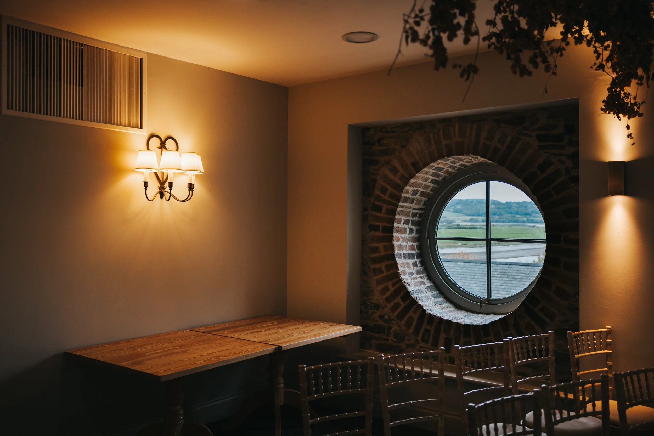 A cozy interior room with a wooden table, a round window framed with bricks, and warm wall lighting.