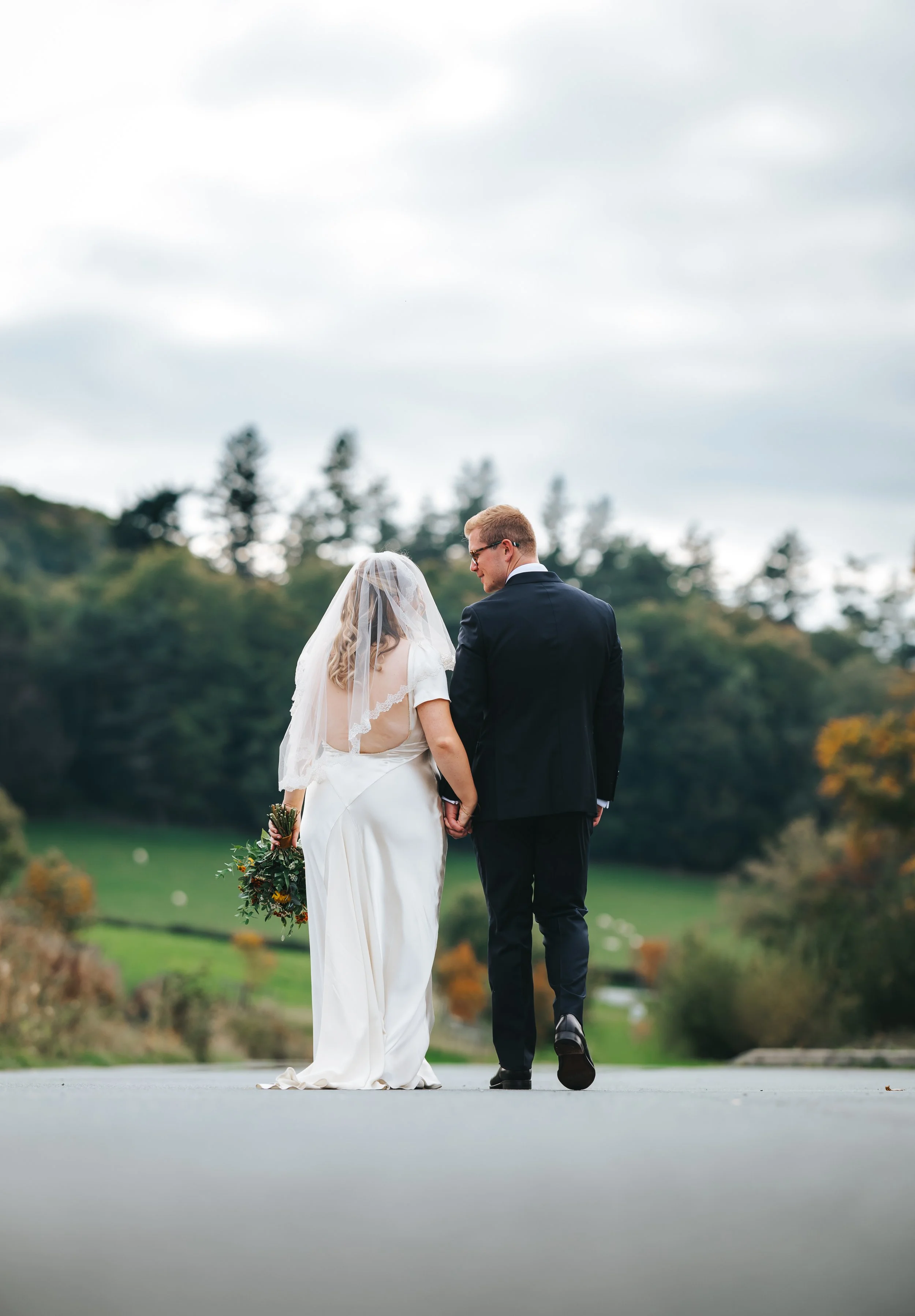 A bride and groom walk hand in hand outdoors during their wedding, with trees and a cloudy sky in the background.