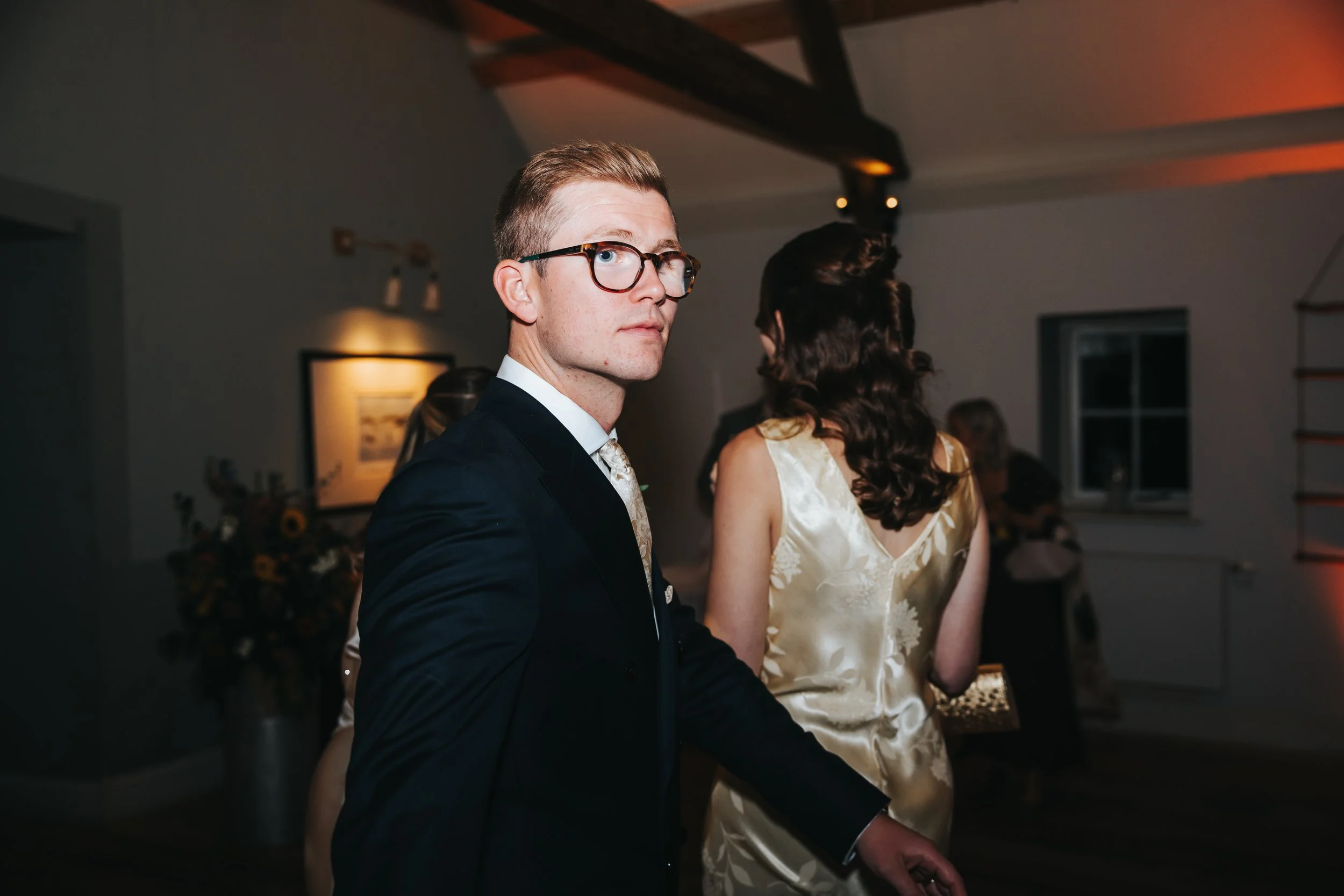 A man in glasses and a dark suit looks over his shoulder at a party or gathering, with a woman in a satin dress in the background.