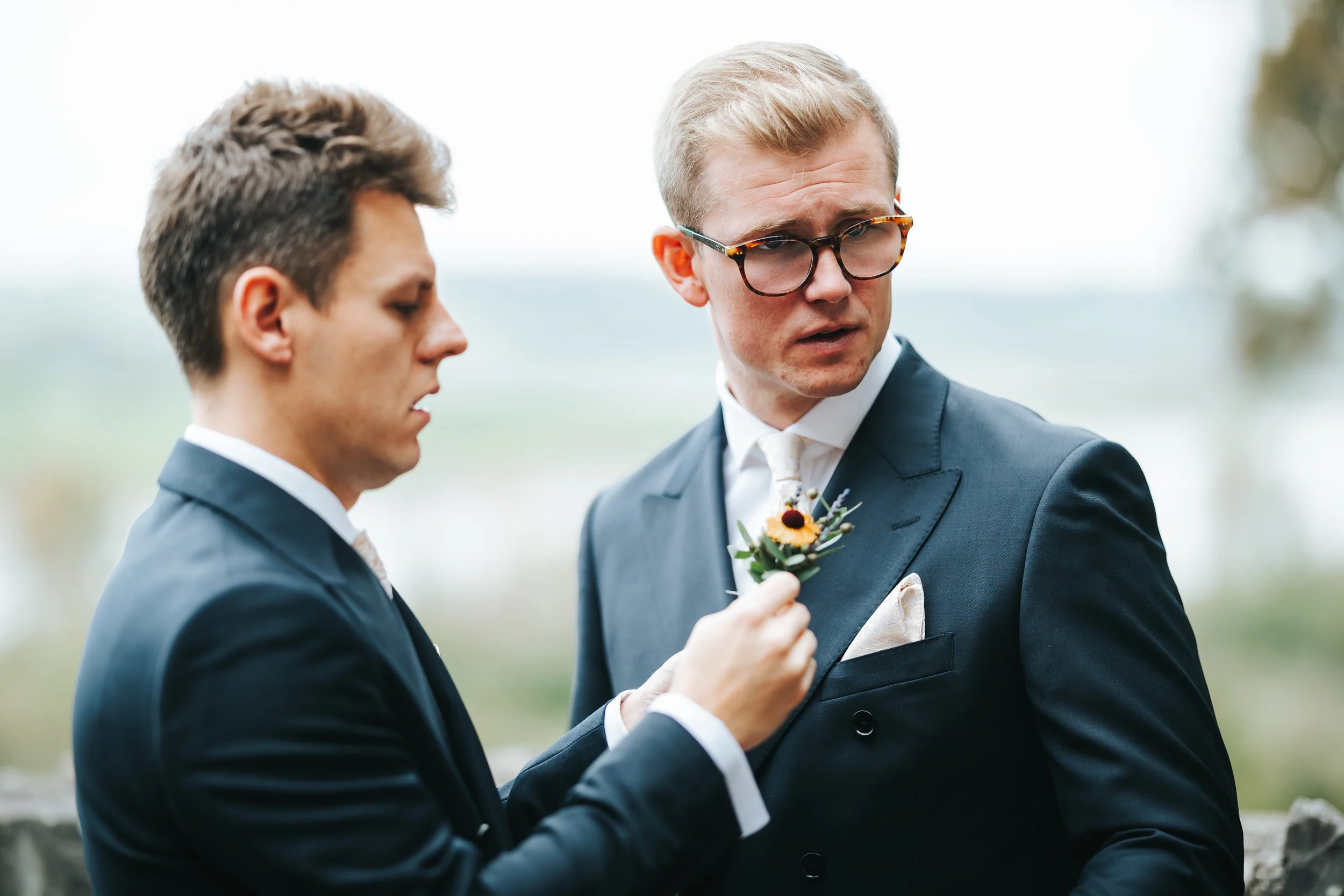 Two men in suits, one pinning a boutonniere on the other's lapel during an outdoor wedding or formal event.