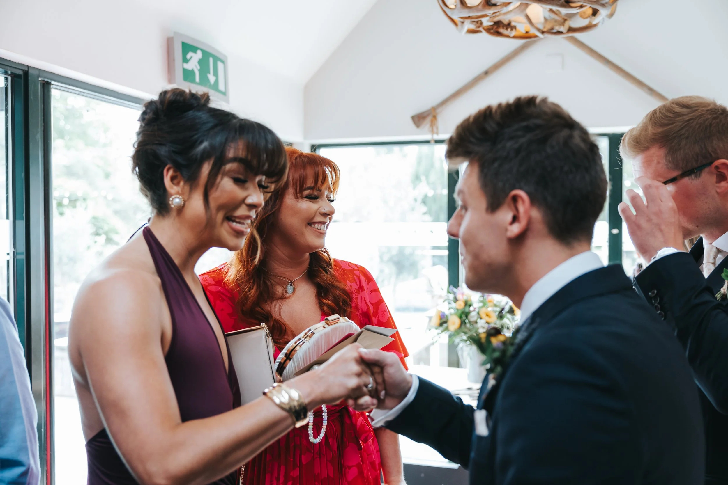 A woman in a purple dress and a woman in a red dress shake hands with a man in a suit during a wedding reception indoors with sunlight coming through windows.