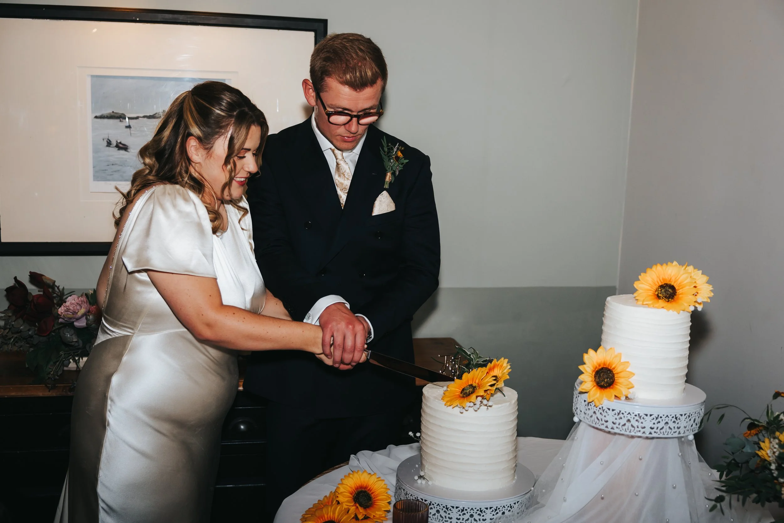 A bride and groom are cutting a wedding cake decorated with yellow sunflowers. They are standing next to a table with a three-tiered cake, and the bride is holding a cake knife with the groom. The bride wears a shiny silk dress, and the groom is in a
