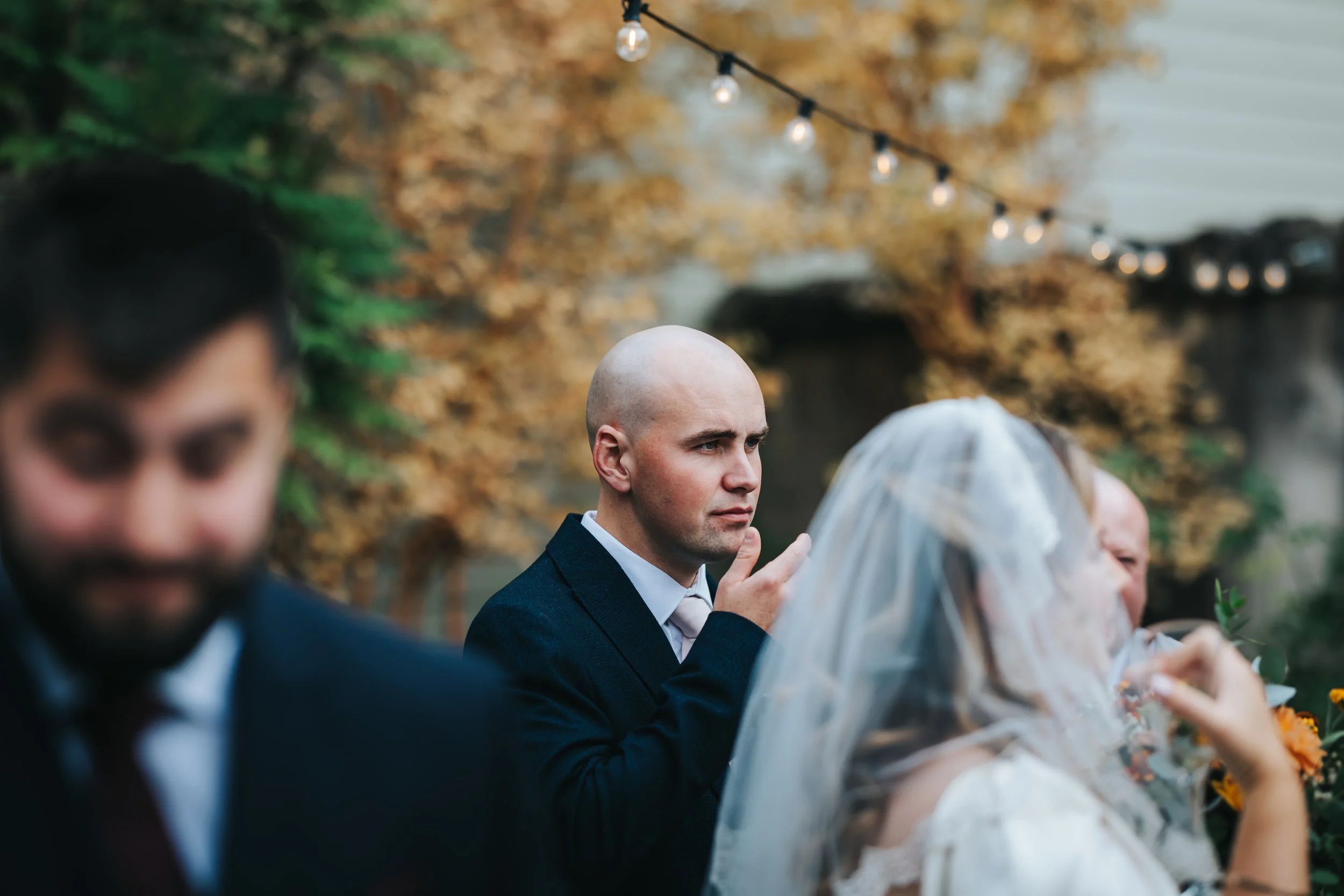 Man with a serious expression at an outdoor wedding ceremony, surrounded by blurred guests and autumn-colored trees, during evening with string lights overhead.